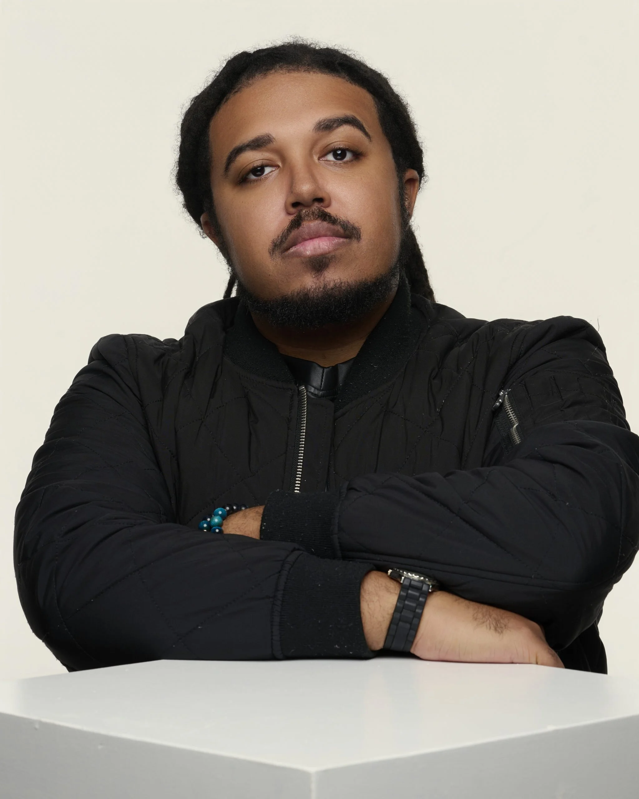 A young man with a beard and dreadlocks sitting with his arms crossed on a white table, wearing a black quilted jacket, a black watch, and a blue beaded bracelet, against a plain white background.