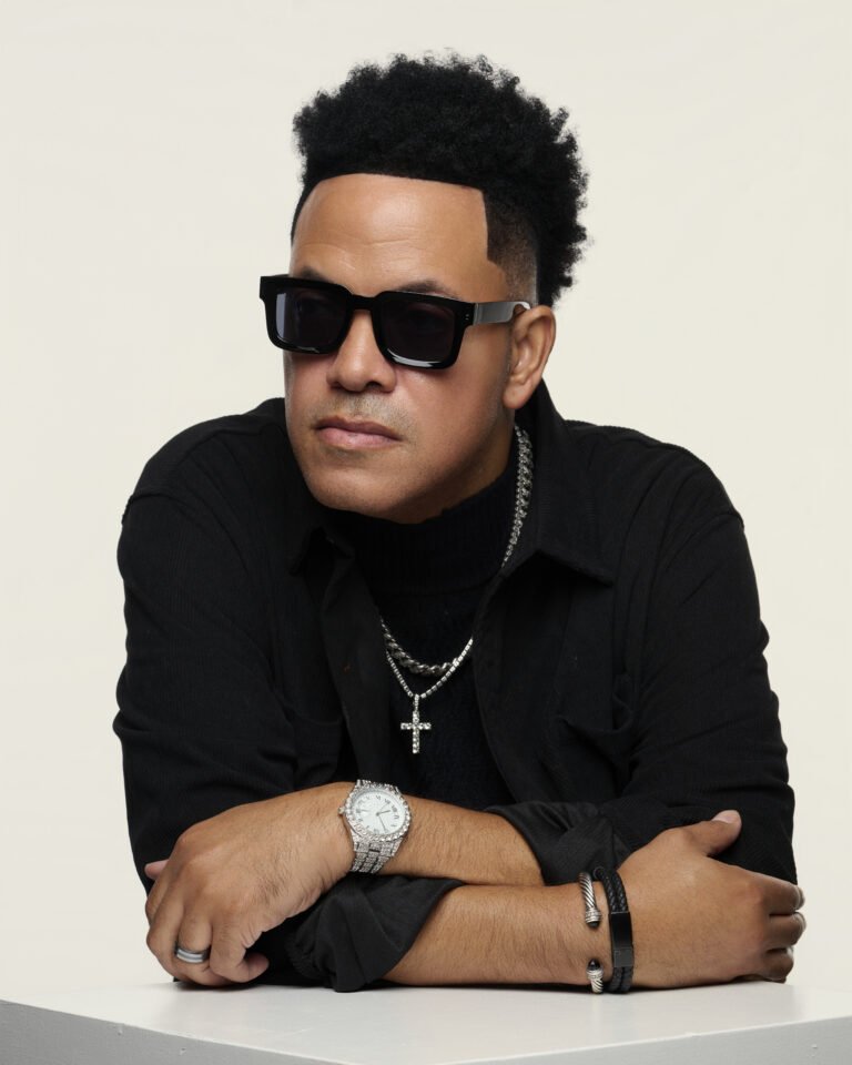 A man wearing black sunglasses, a black shirt, and jewelry including a chain with a cross, a watch, and bracelets, poses with his arms crossed on a white surface against a plain background.