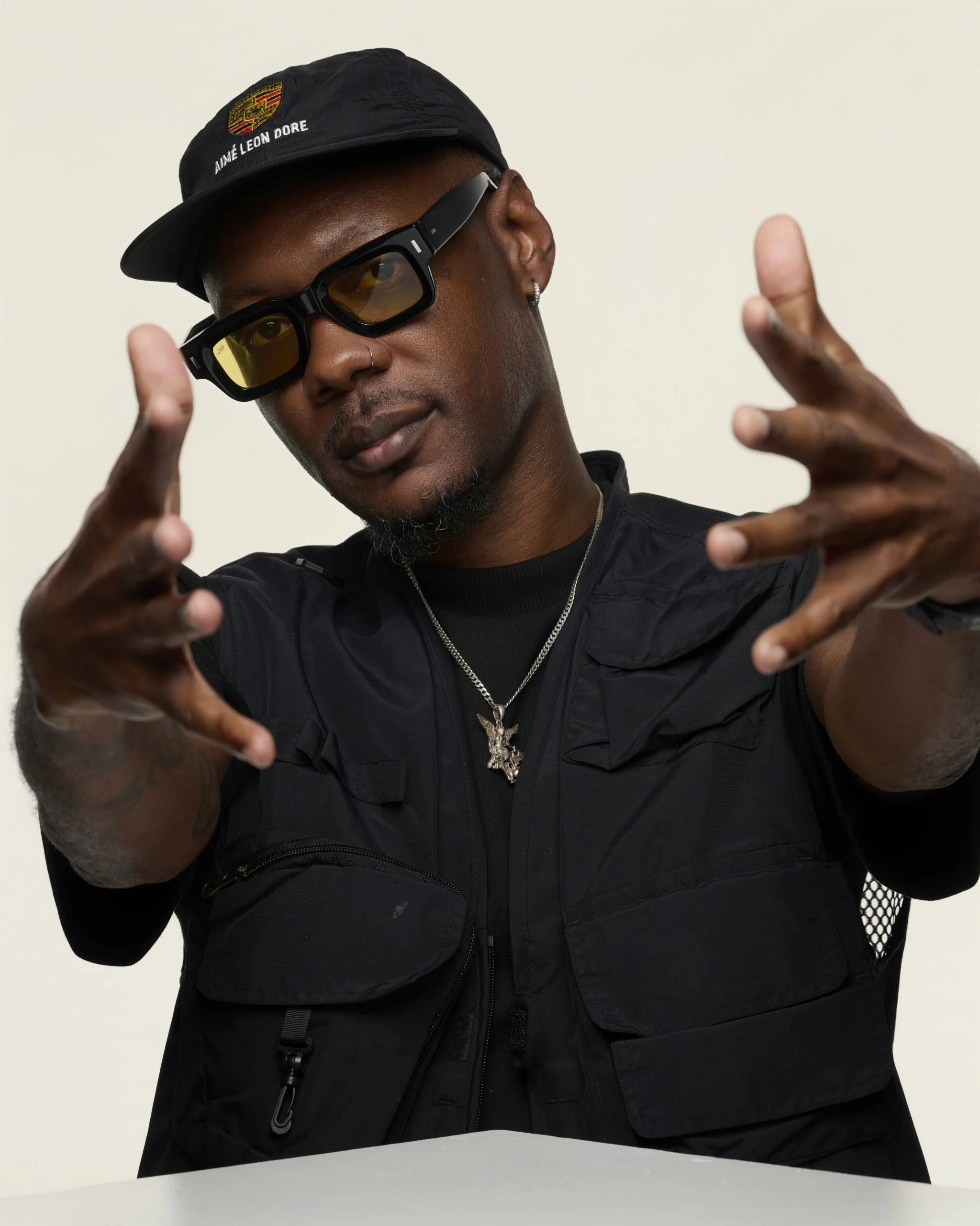 A young man wearing a black cap with a Porsche logo and the words 'Aimé Léon Dore', black sunglasses with yellow lenses, multiple earrings, a nose ring, and a silver chain with a pendant, making a peace sign gesture with both hands in front of a plain background.