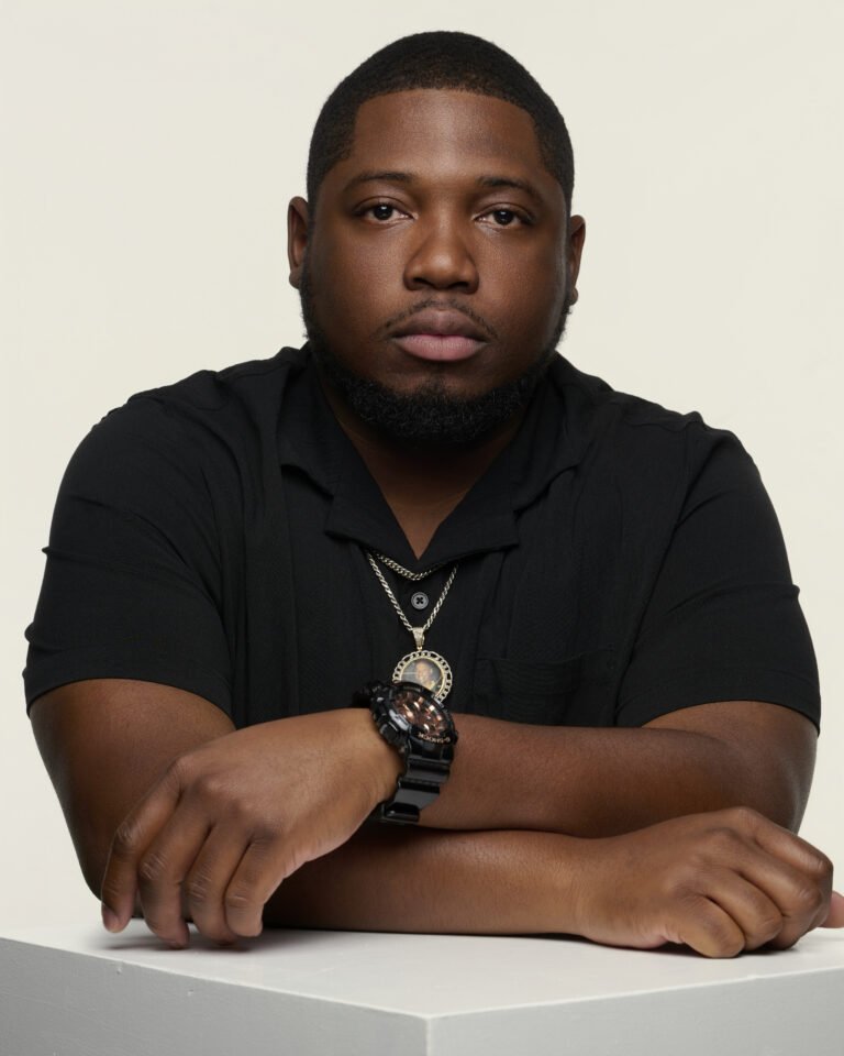 Portrait of a young man with short black hair and beard, wearing a black shirt, watch, and necklace, posing with arms crossed in front of a plain light background.