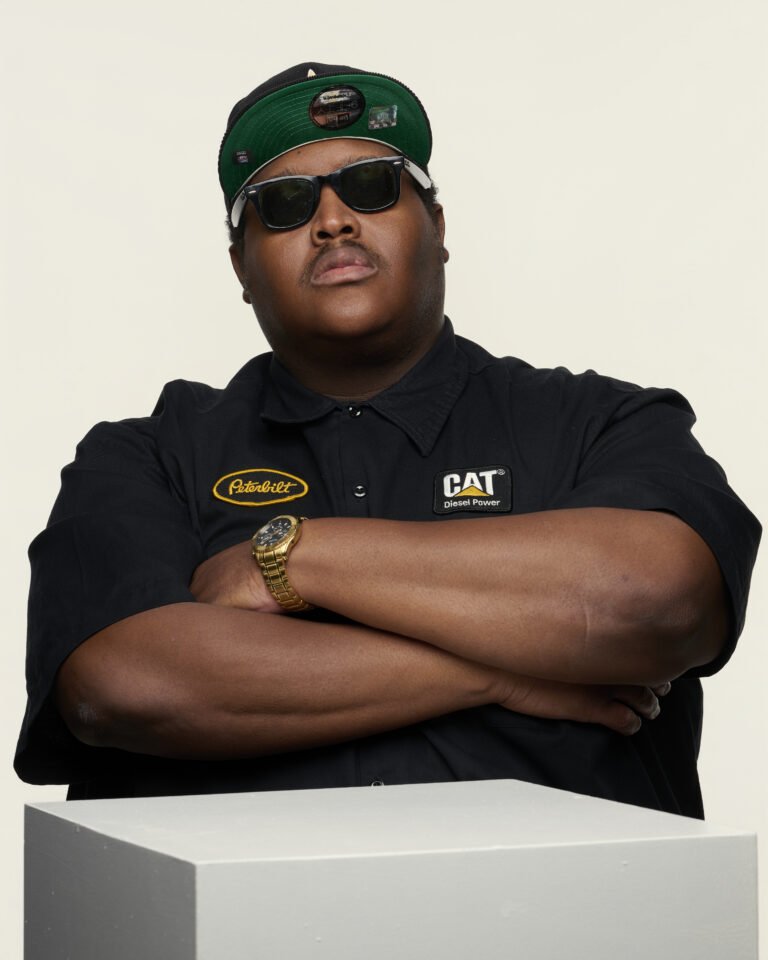 A man wearing black sunglasses, a black cap with a green brim, and a black shirt with CAT Diesel Power and Paterbilt patches, crossing his arms, standing behind a white surface.