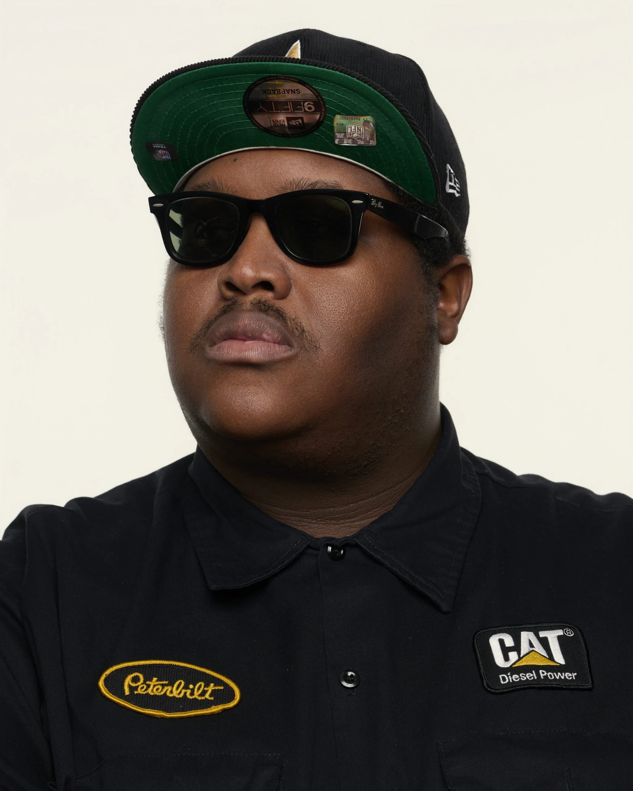 Close-up of a person wearing a black cap with a green underside, black sunglasses, and a black jacket with patches. The patches display logos for Caterpillar and Petro-Lift.