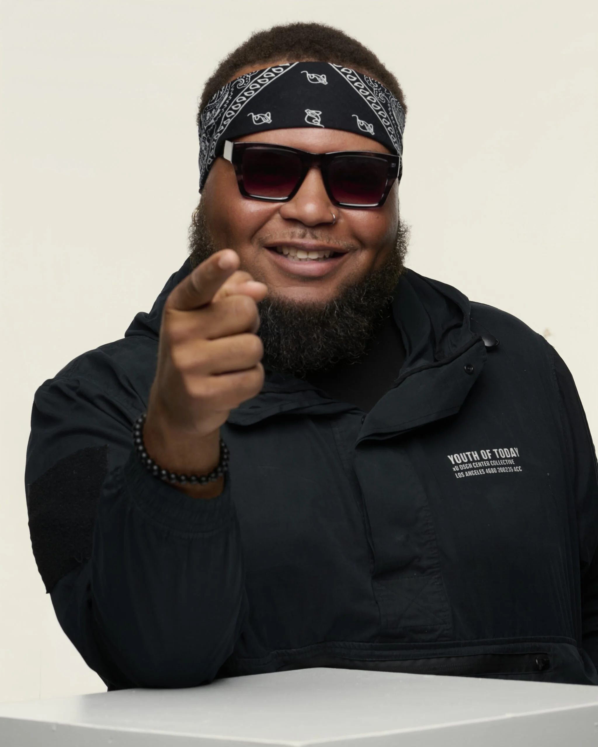 Smiling man with beard and nose piercing, wearing large sunglasses and a black bandana, pointing towards the camera, dressed in a black jacket with white text on the chest, sitting at a white table against a plain light background.