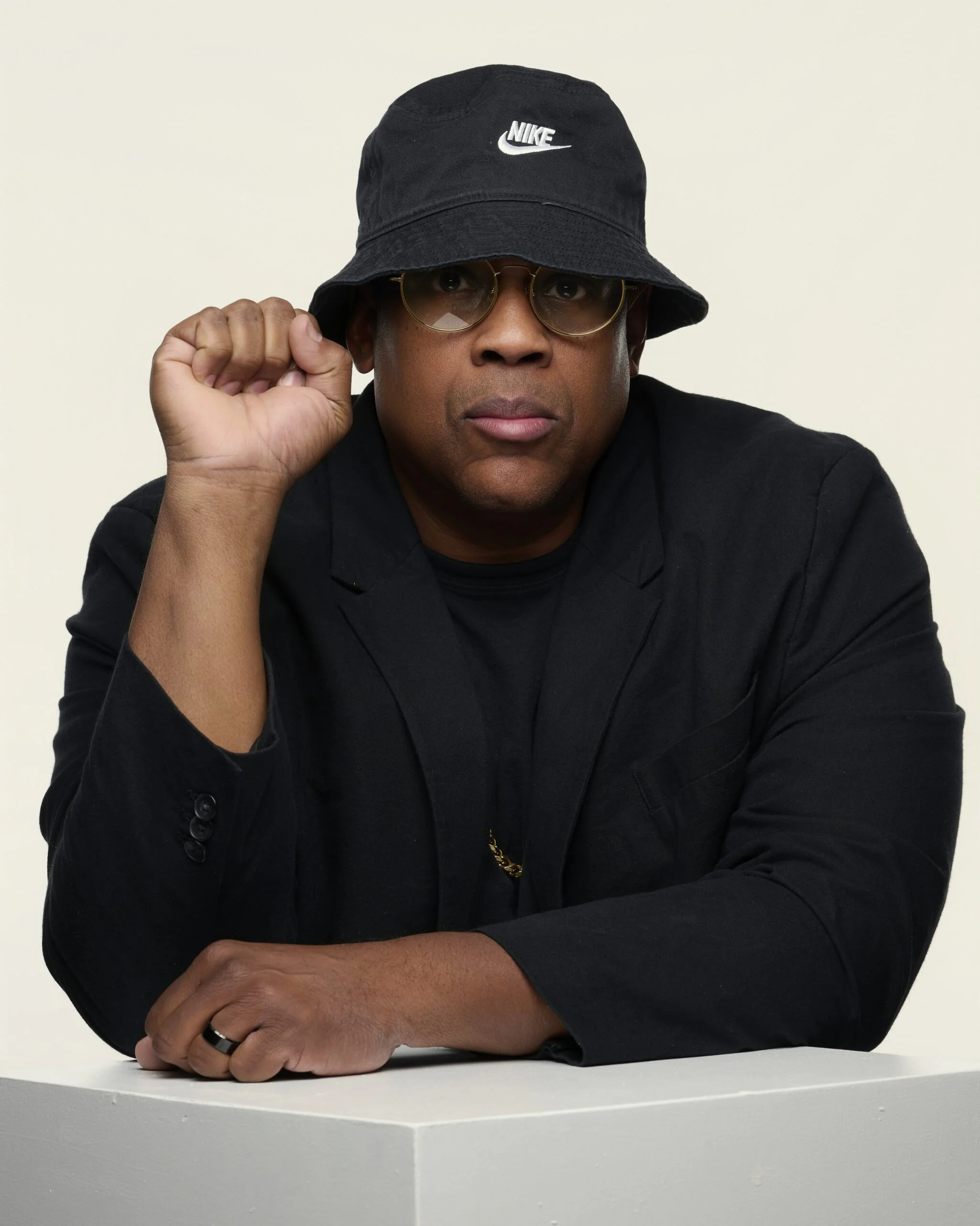 A man wearing gold-rimmed glasses and a black Nike bucket hat, sitting at a white table against a plain white background, with his right hand adjusting his glasses and his left arm resting on the table.