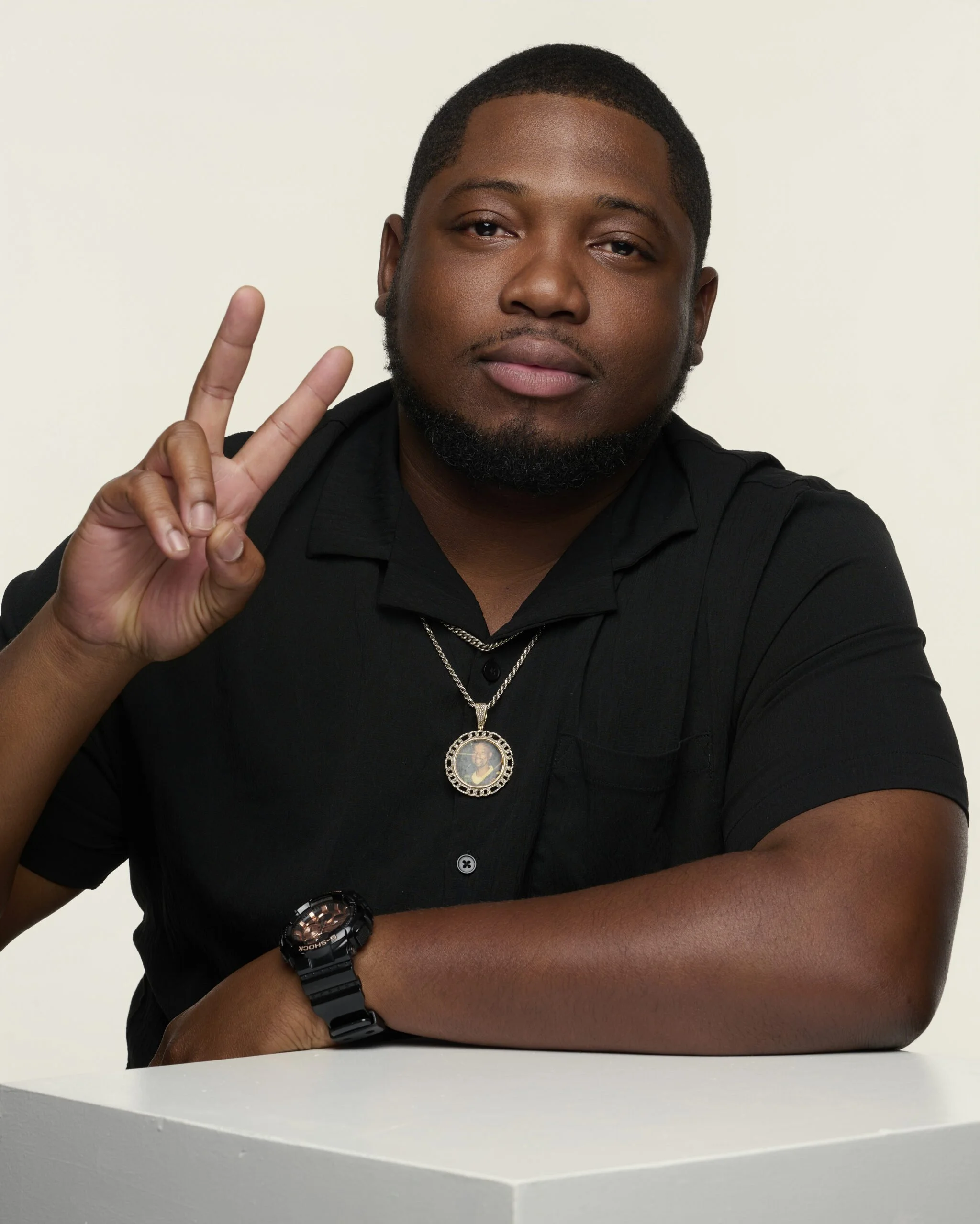 A young man with short hair and a beard making a peace sign with his right hand. He is wearing a black shirt, a black wristwatch, and a necklace with a framed photo pendant. He is sitting at a white table against a plain white background.