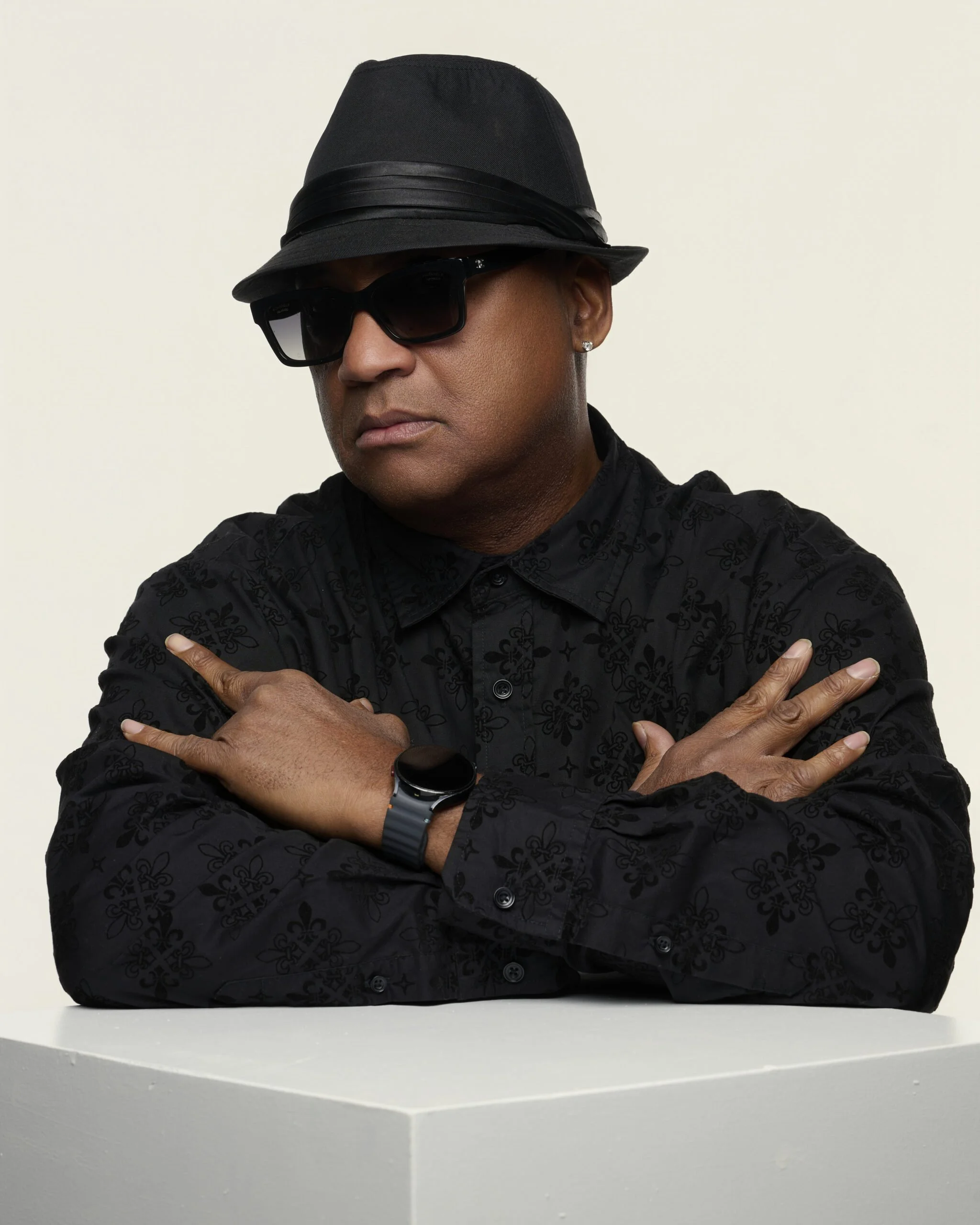 A man wearing a black fedora hat, black sunglasses, and earrings, crossing his arms while standing against a white background.