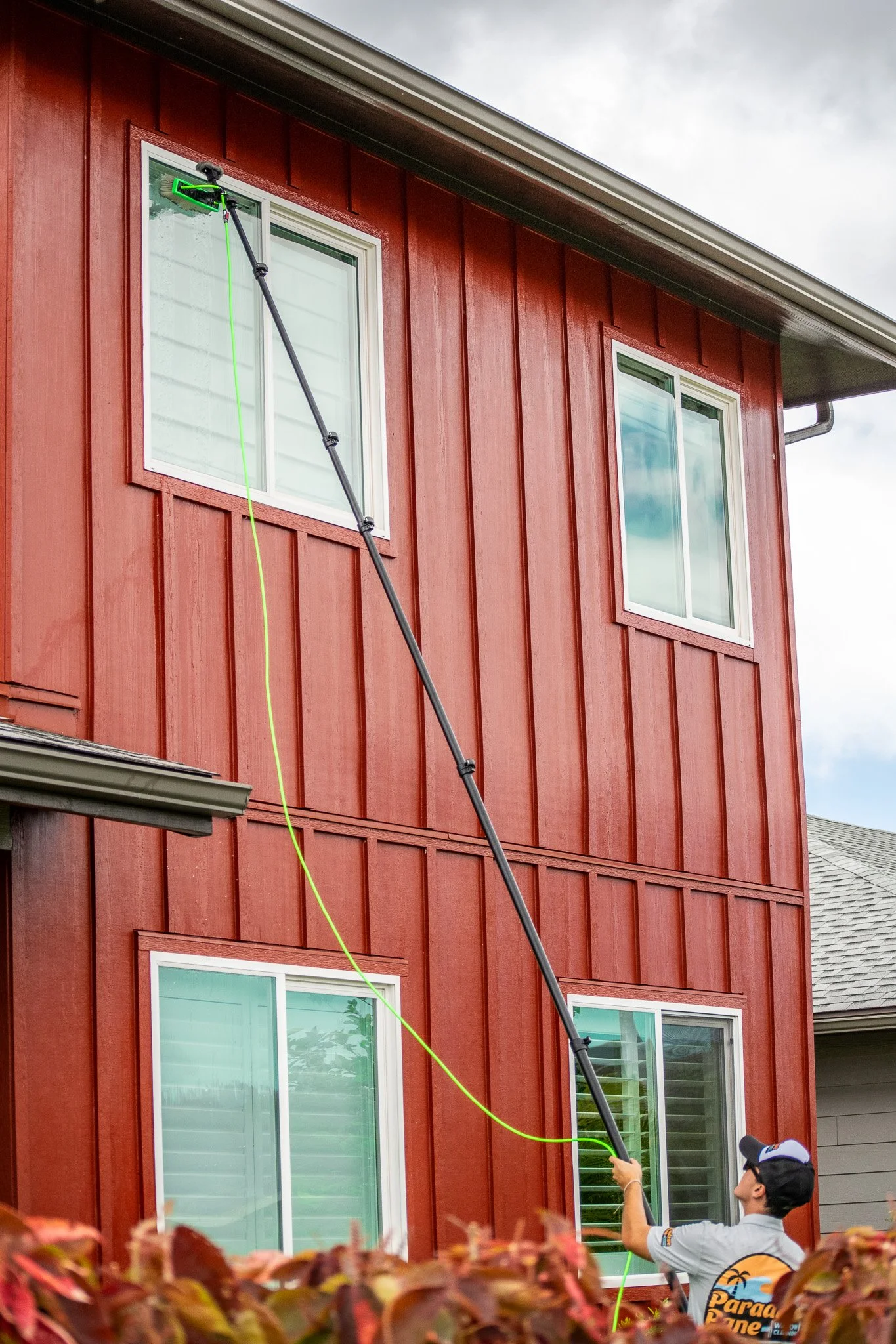 A person cleaning the exterior of a red house window with a long pole and a spray brush, wearing a hat and a shirt with the words "Para Gone" on the back.