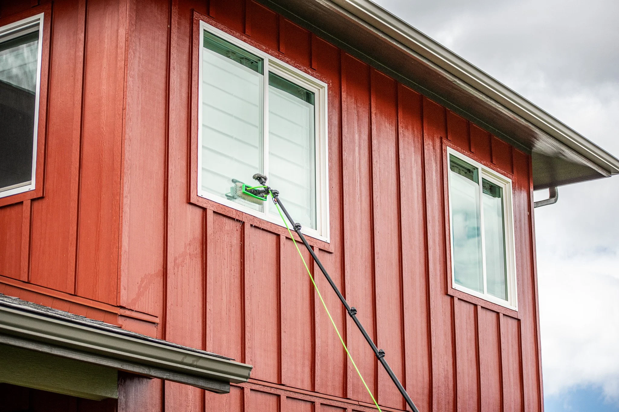 A red house with tall vertical siding and two windows, one with a lime green and black tool resting on the window ledge, an extension pole leaning against the house, and a cloudy sky in the background.