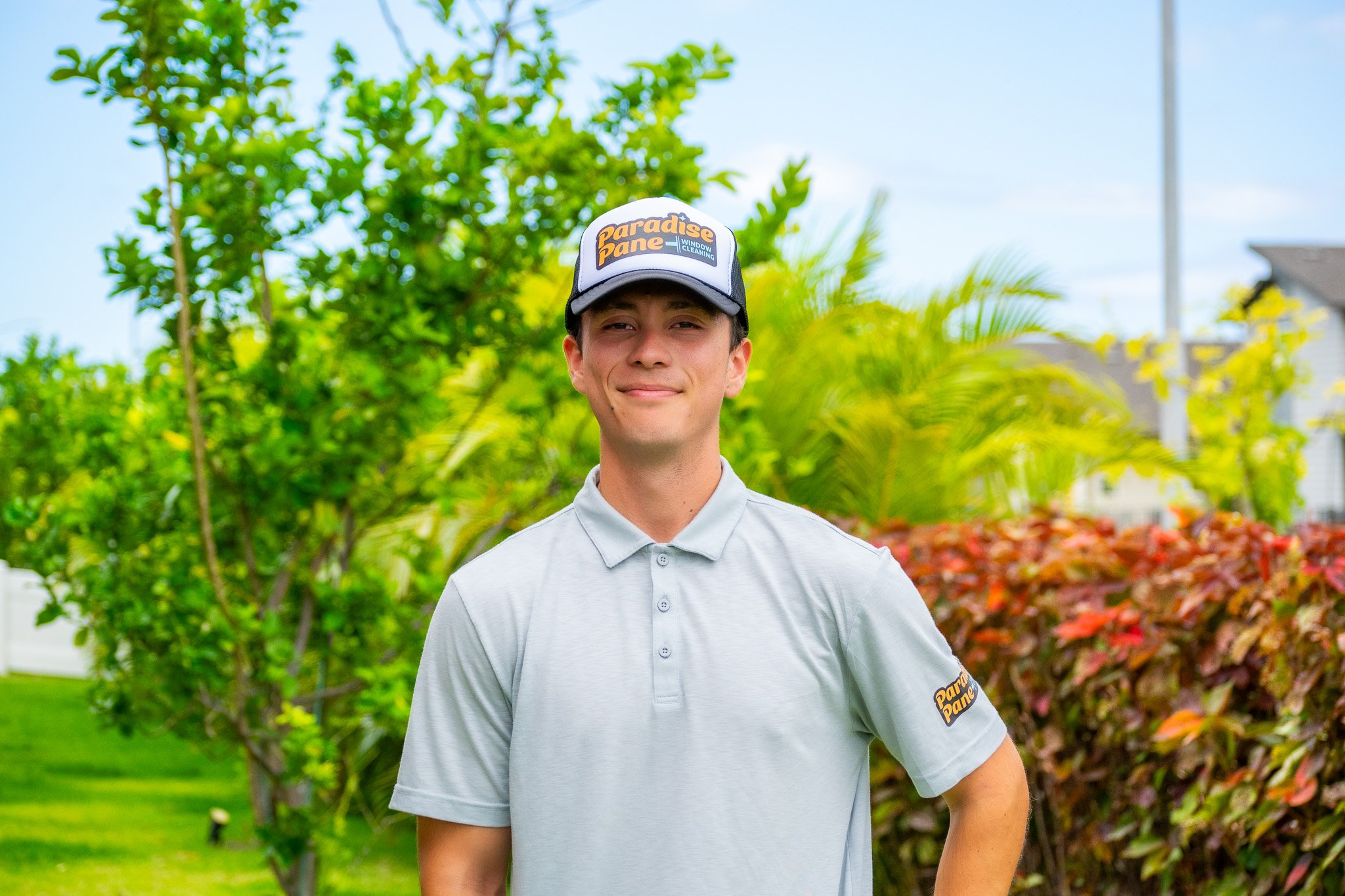 A young man in a light gray polo shirt and a black and white cap with the logo 'Paradise Pane Window Cleaning' stands outdoors with lush green trees and colorful shrubs behind him.