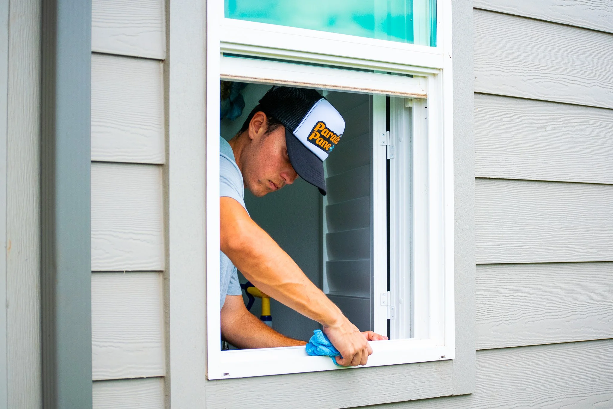A man wearing a black baseball cap with the words "Paradise Pane Window Cleaning" visible, leaning out of a house window cleaning the exterior with a blue cloth.