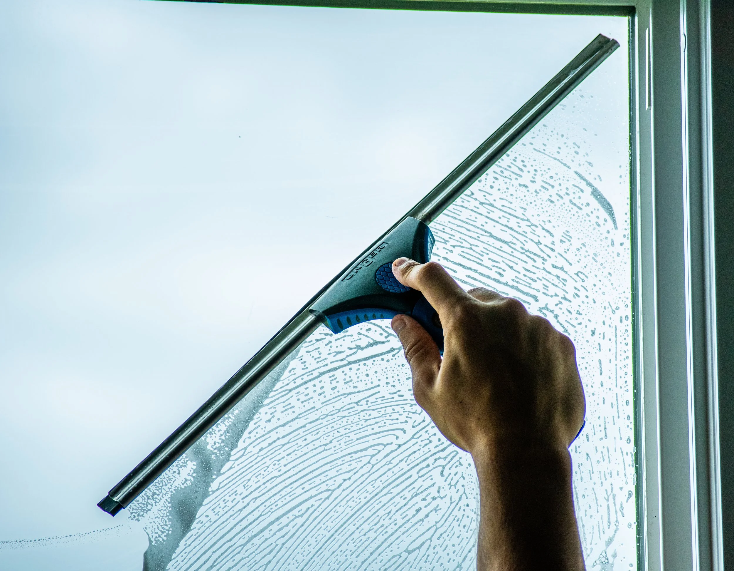 A person using a long-handled squeegee to clean a glass window, with soap or cleaning solution visible on the glass.