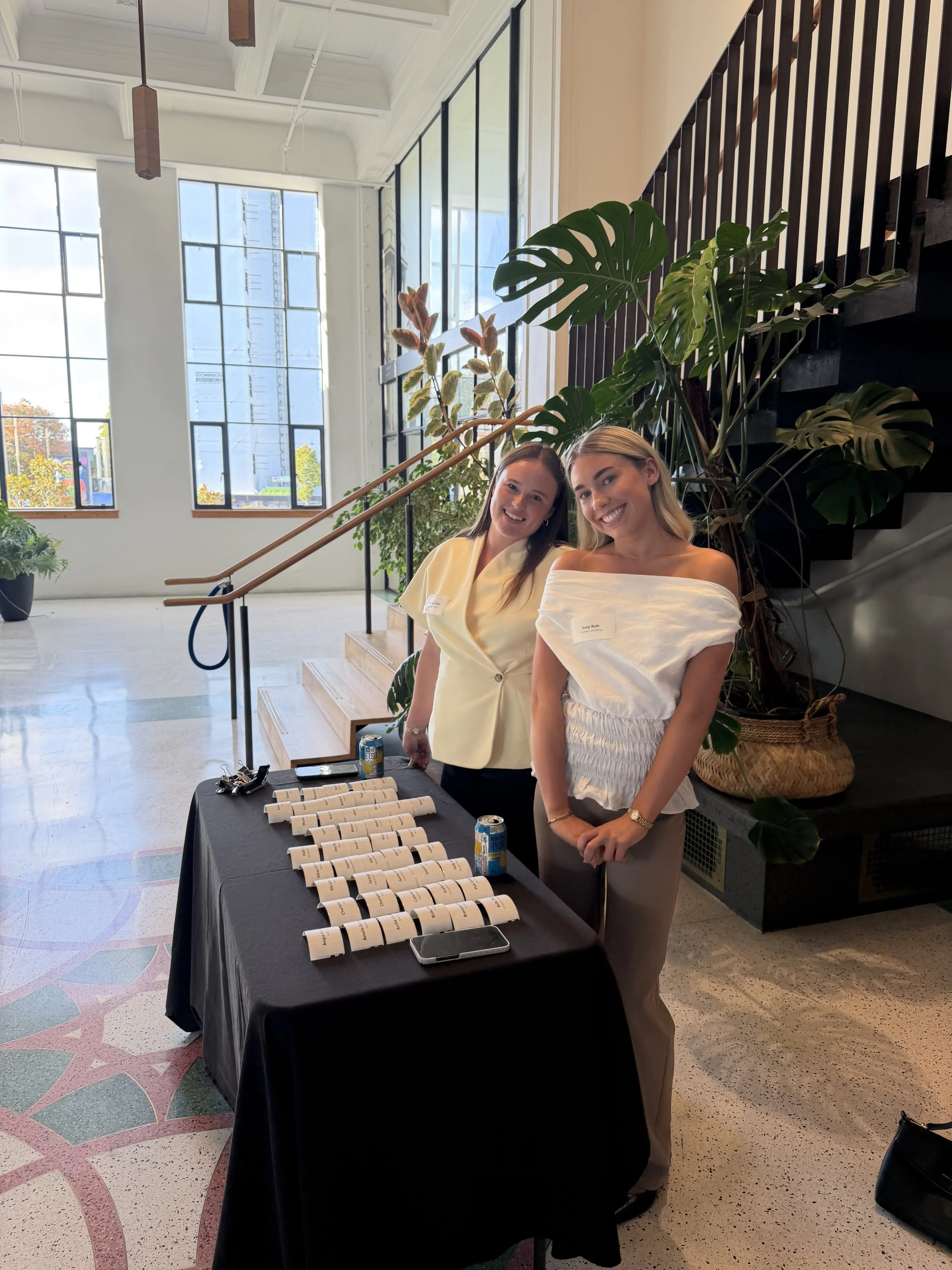 Two women standing behind a table with small rolled papers and drinks in an indoor space with large windows, plants, and staircase in the background.