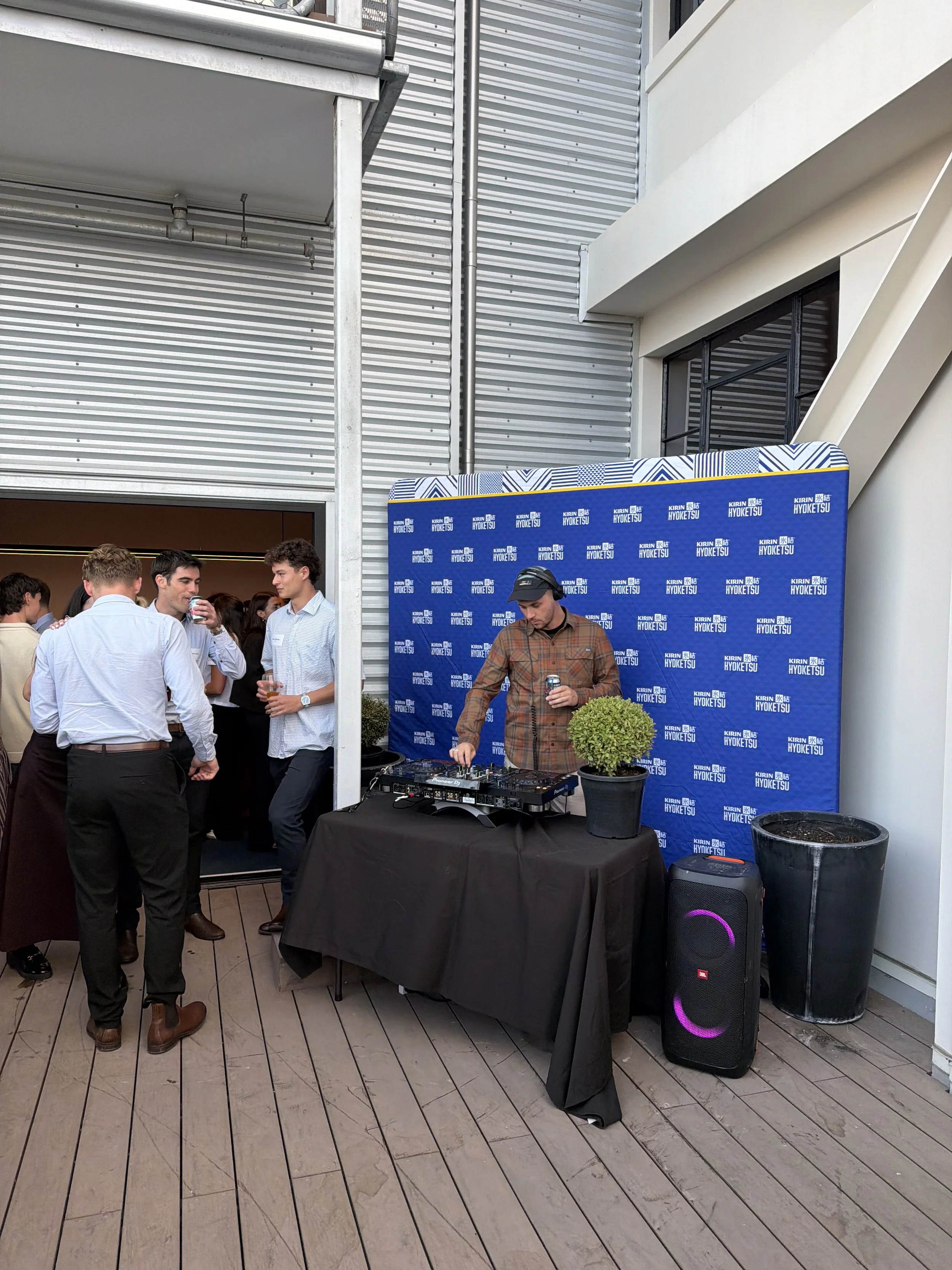 People gathered in an outdoor space with a DJ performing at a setup with speakers and a backdrop with the text "KIRIN HYOKETSU." Some individuals are socializing, and a few are drinking beverages. The area has a wooden deck floor and modern building architecture.