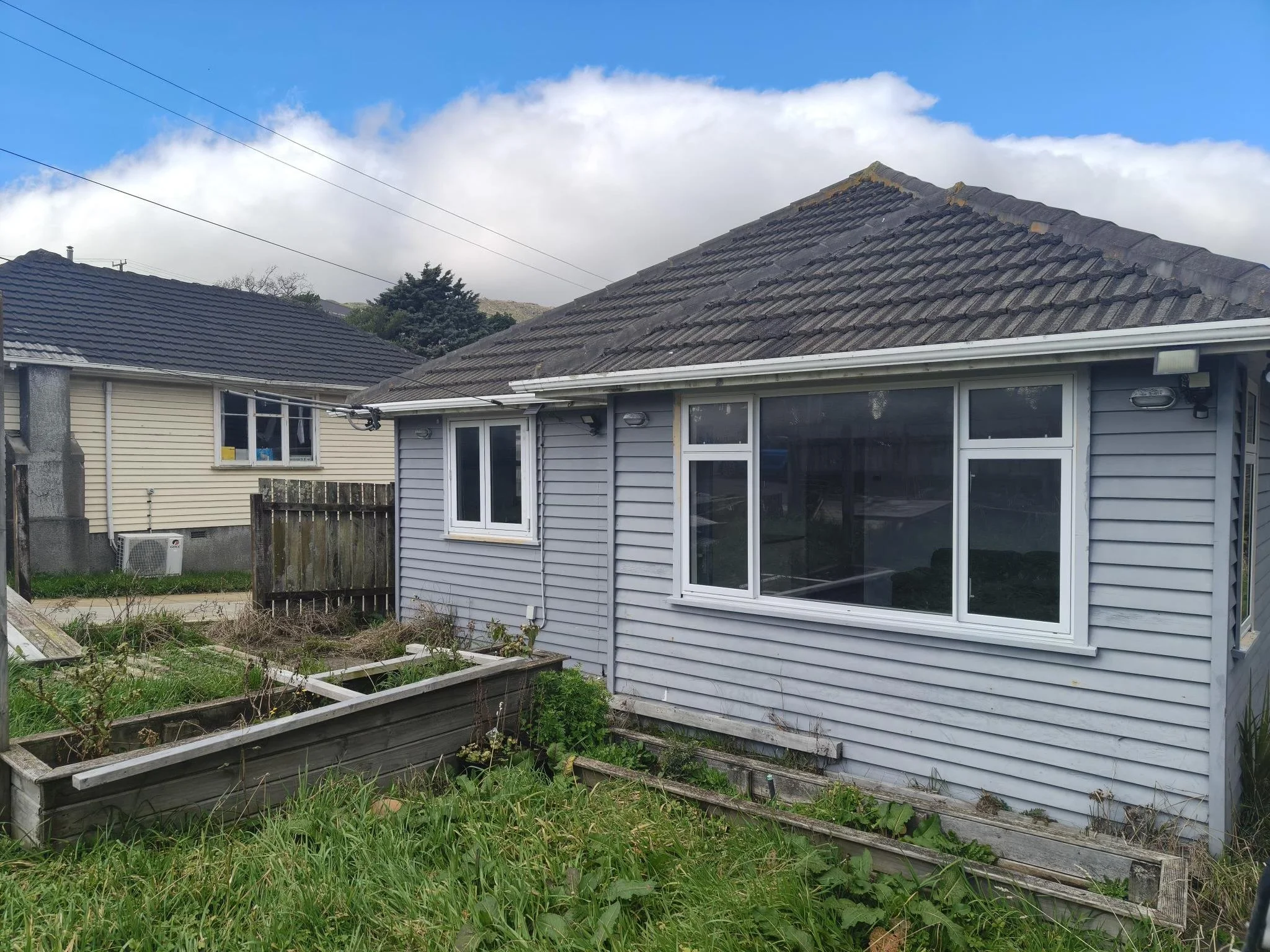 A backyard view of two houses with different roof styles, a garden bed in the foreground, and a partly cloudy sky.