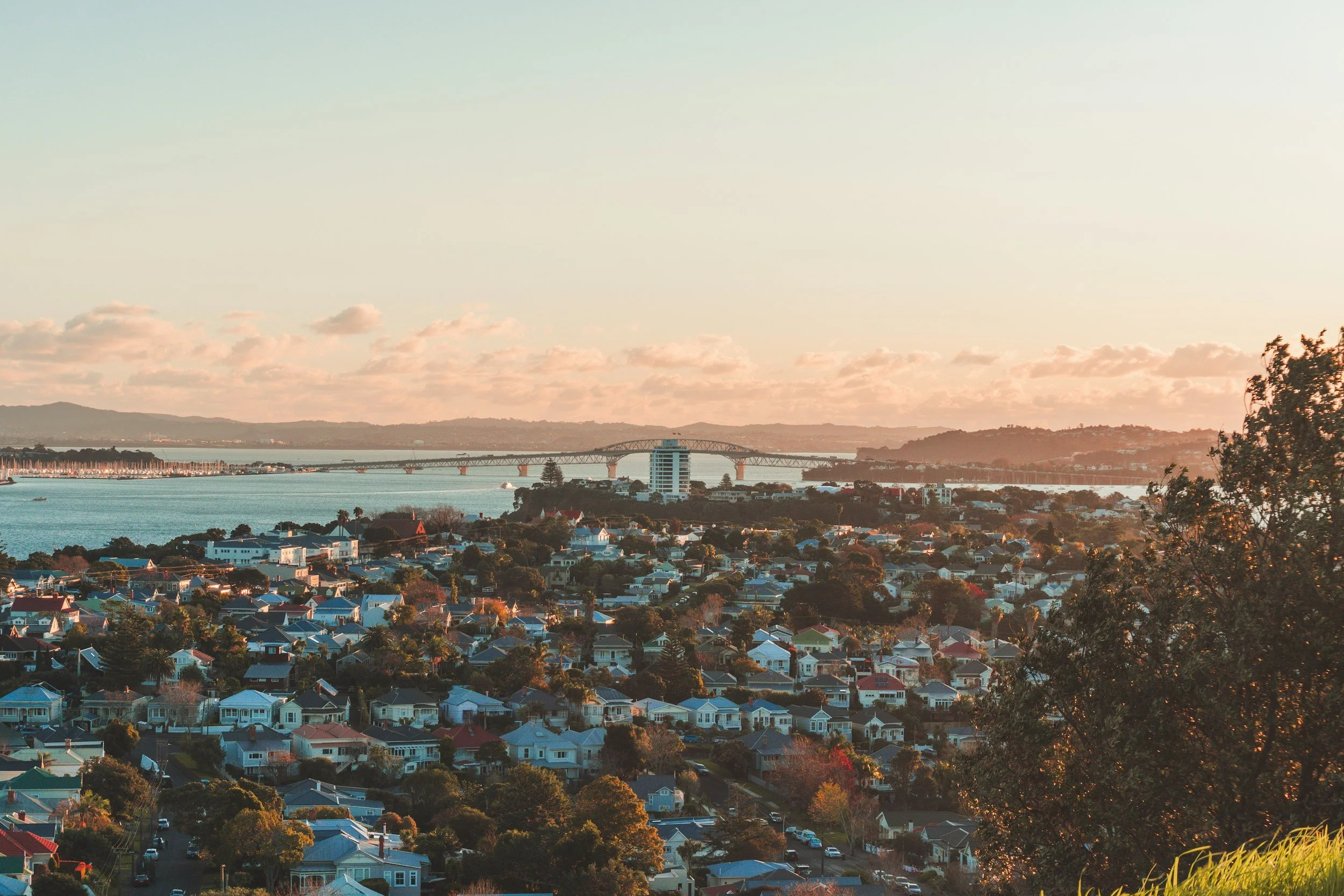 A cityscape with houses in the foreground, a body of water, a bridge, and hills in the distance during sunset.