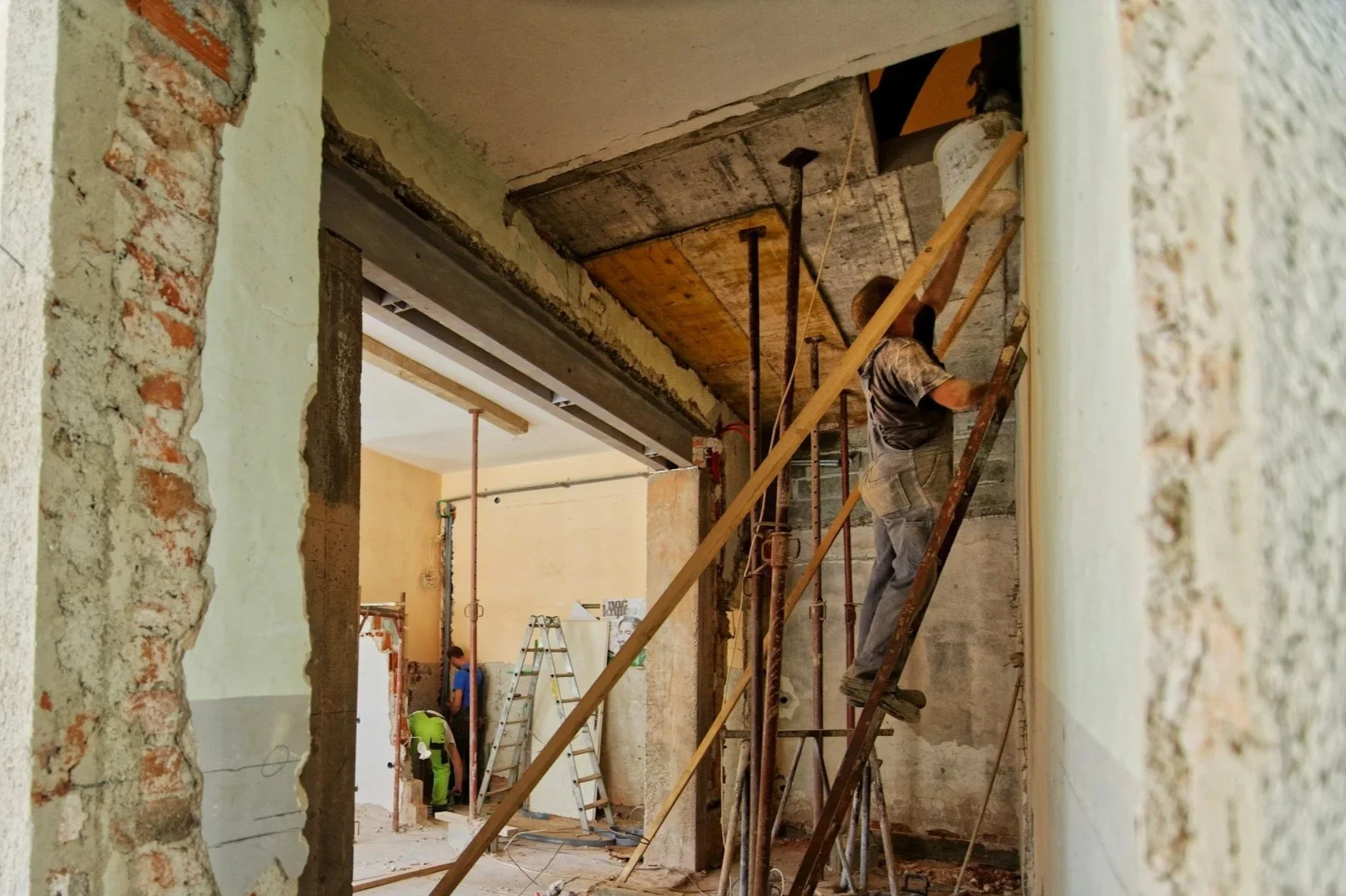 Construction workers renovating an interior space with exposed brick, concrete, and wood elements. One worker is on a ladder, working near the ceiling, while others are visible in the background.