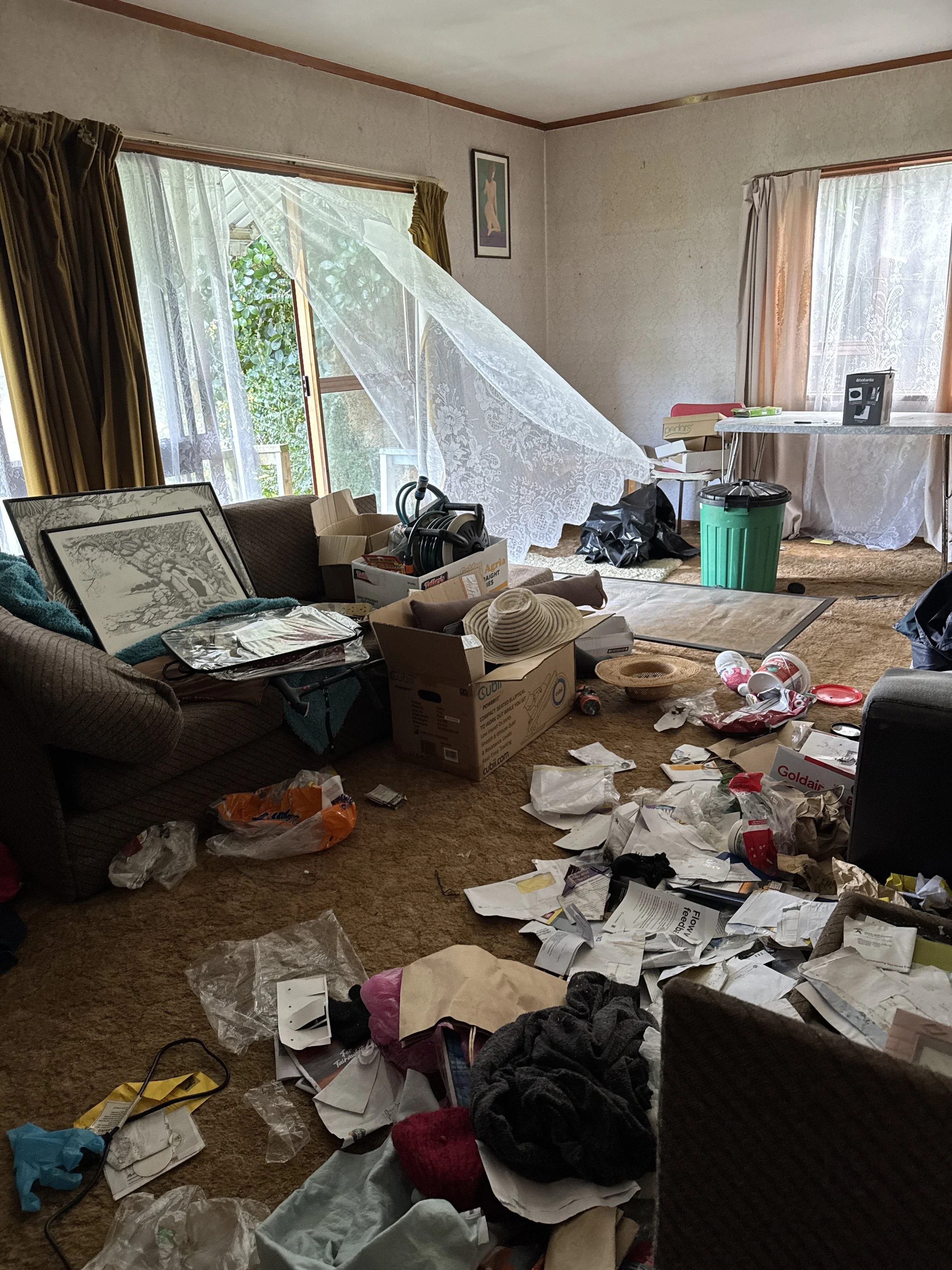 A cluttered living room with scattered trash, papers, and household items on the floor and furniture, including a couch, table, and window with curtains.