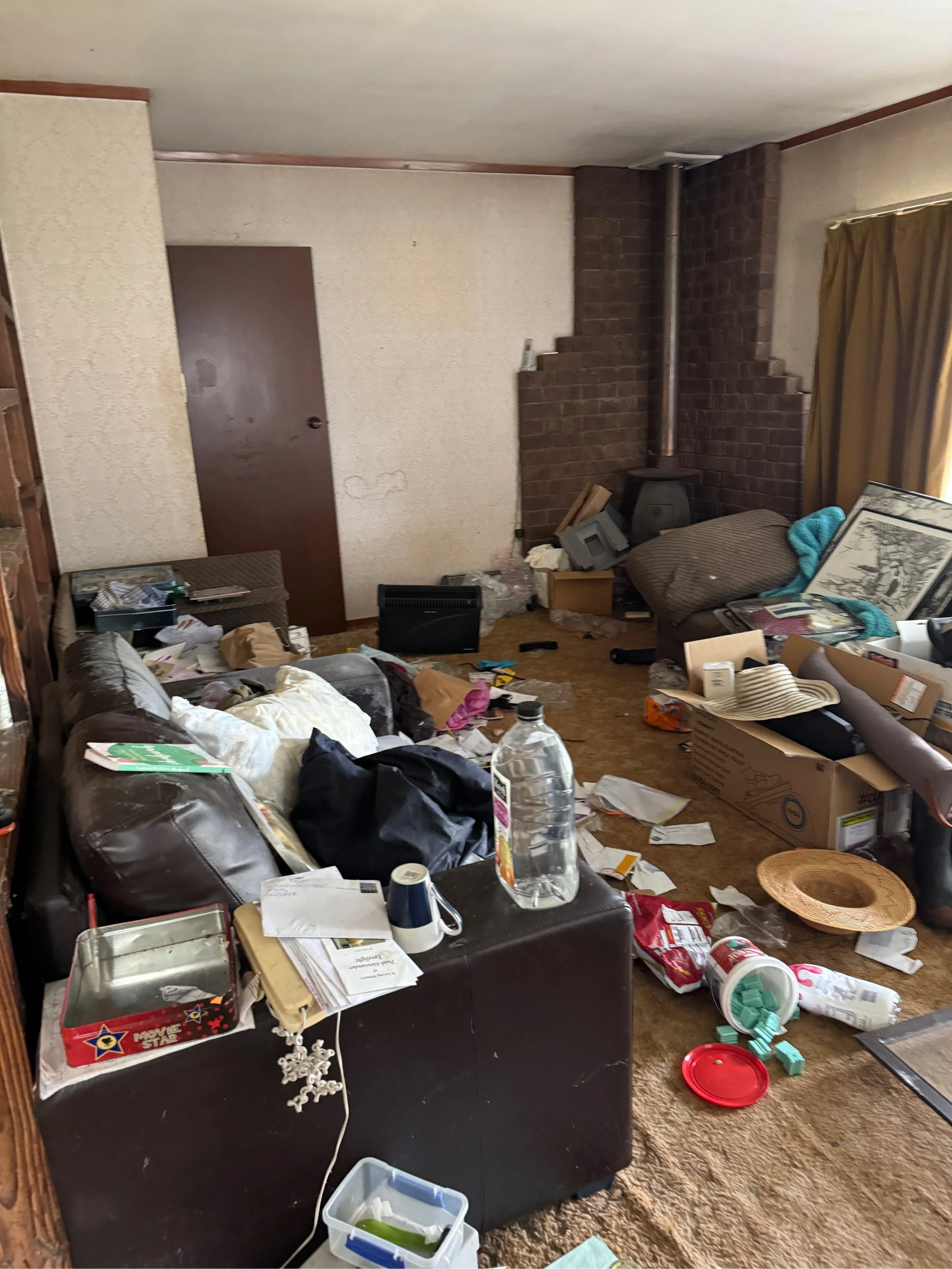 A cluttered living room with a black couch, coffee table, and a pack of cigarettes. The room has stained carpet, a brick fireplace, and a door on the wall. The floor and furniture are covered with trash and various items including cups, papers, and b