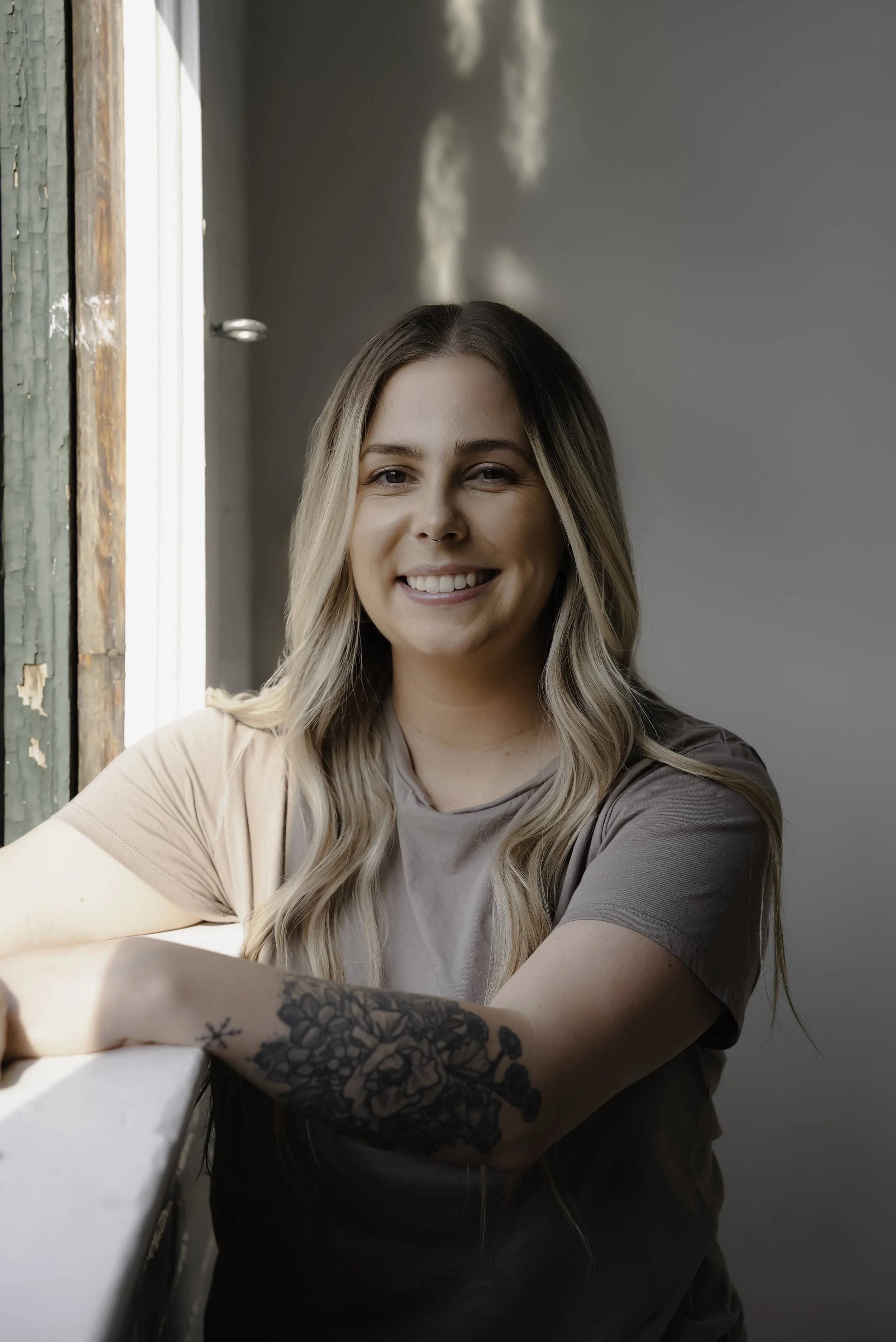 A woman with long blonde hair smiling, sitting near a window with sunlight casting shadows on the wall behind her. She is wearing a short-sleeved shirt, and has a tattoo on her forearm.