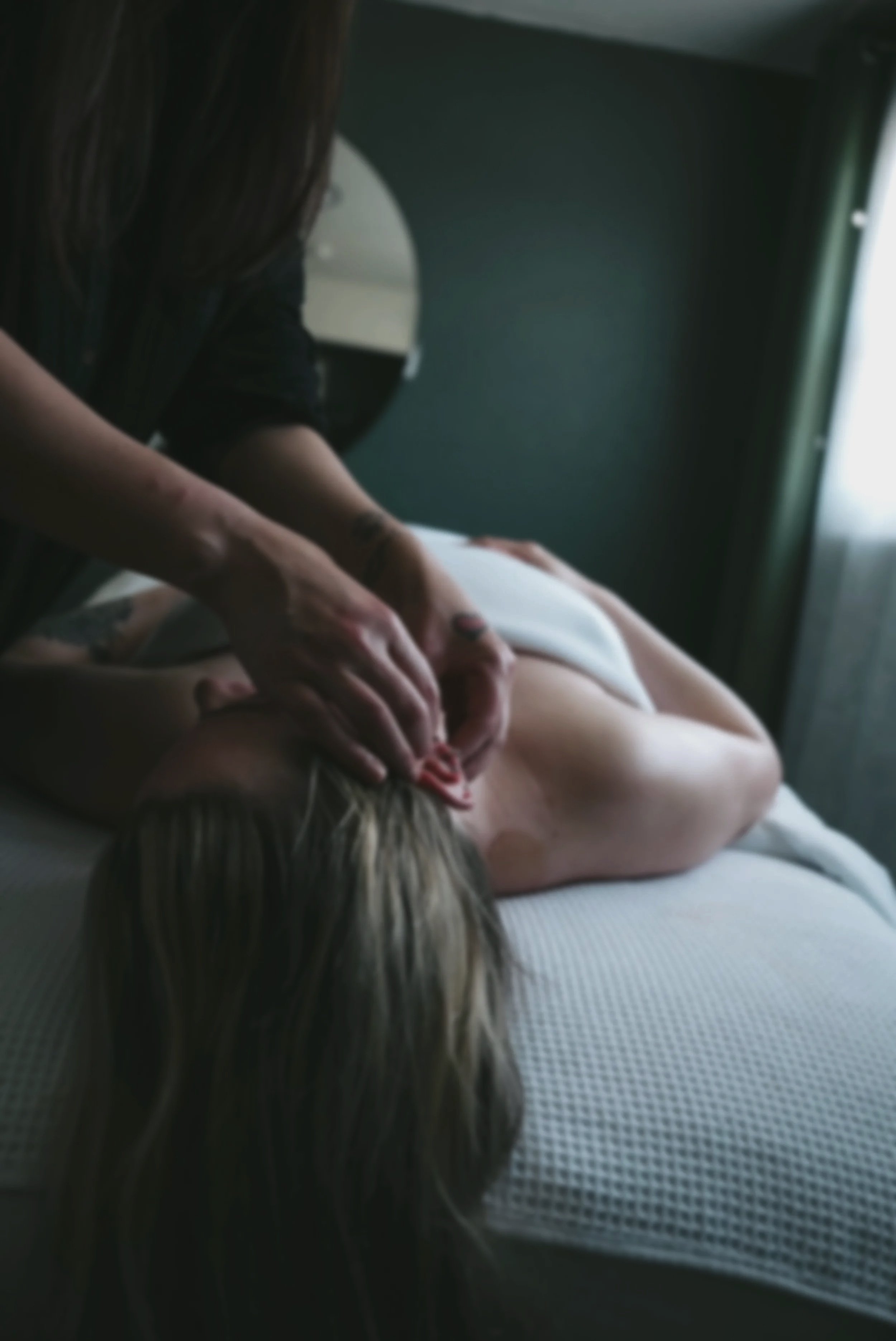 A person giving a massage to another person lying on a massage table in a dimly lit room, with a mirror reflecting part of their bodies.