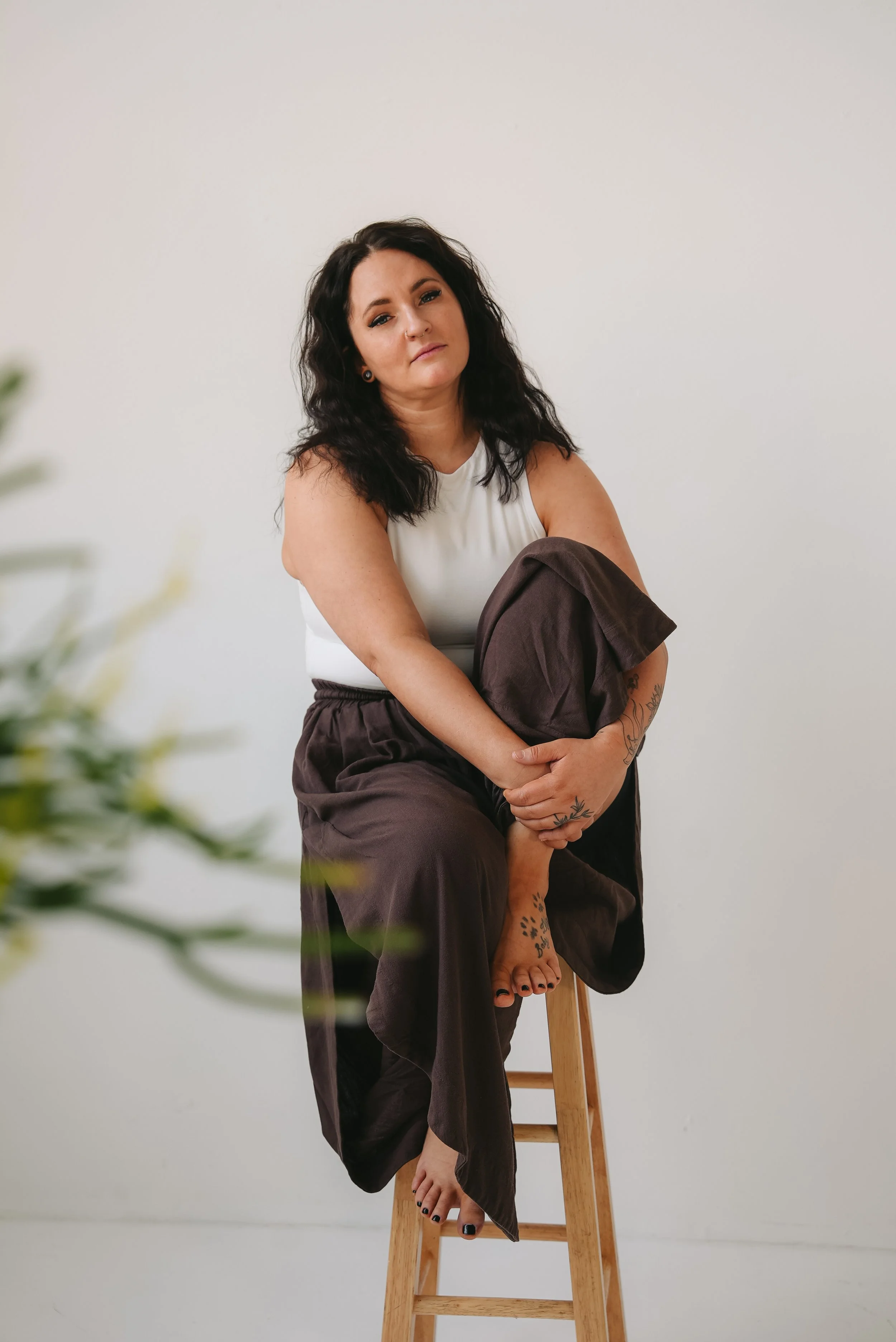 A woman with dark wavy hair and tattoos on her foot and arm, sitting on a stool with one leg crossed over the other, holding a pair of dark pants. She is wearing a white sleeveless top and has a neutral expression, against a plain white background.