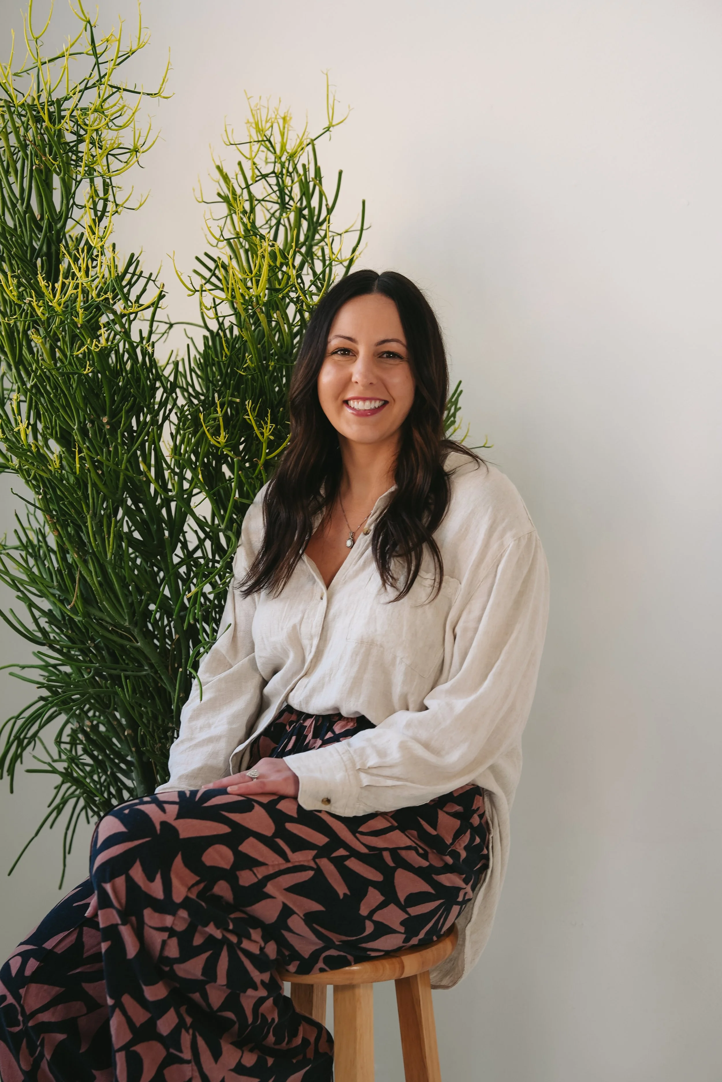 A woman with long dark wavy hair and a big smile, wearing a cream-colored shirt, colorful patterned pants, sitting on a wooden stool beside a tall green plant with thin, yellow-tipped stems, against a plain white wall.