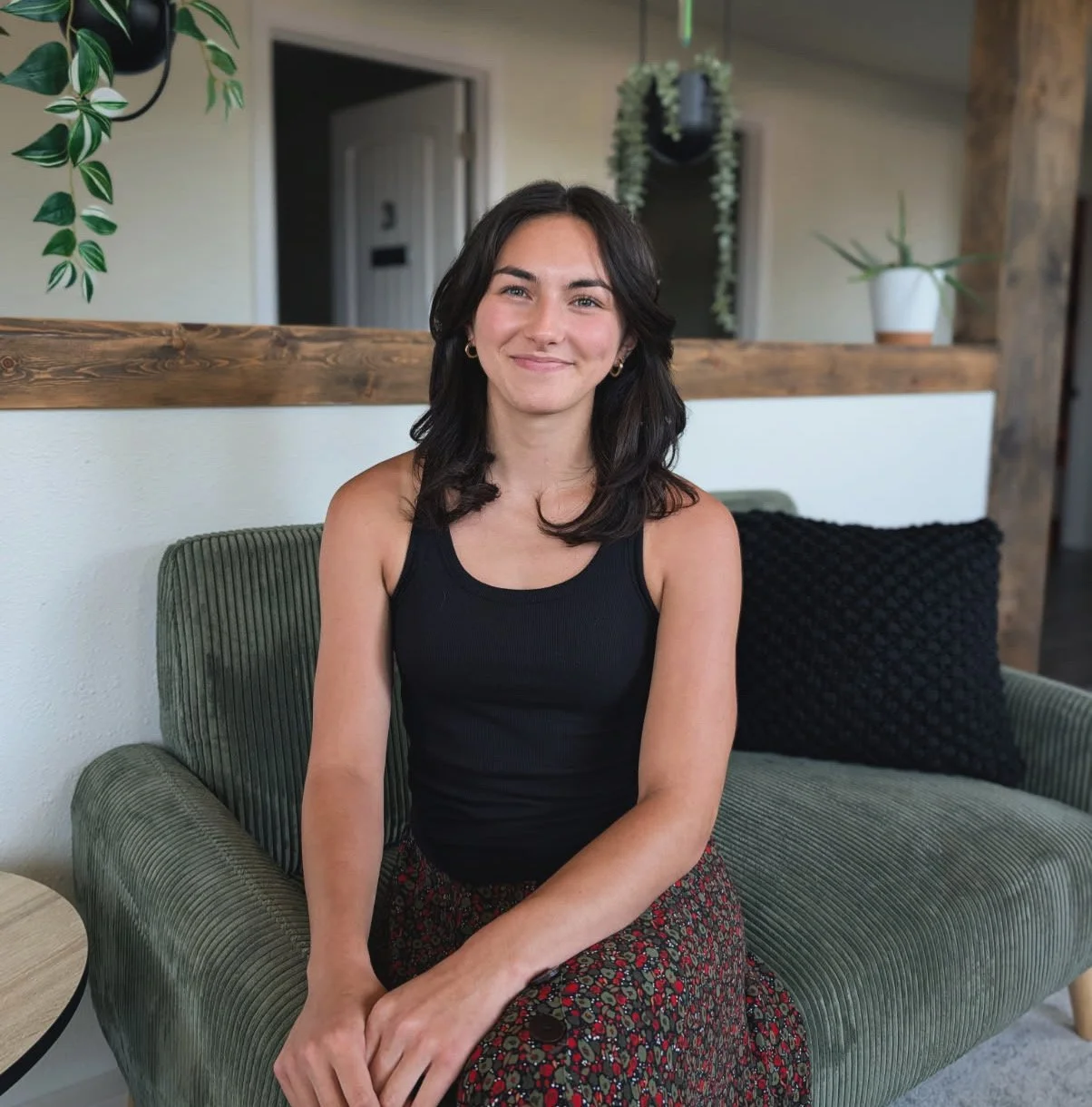 A young woman with dark hair and light skin, smiling while sitting on a green velvet sofa in a cozy interior. She is wearing a black tank top and a patterned skirt, with indoor plants and wooden accents in the background.