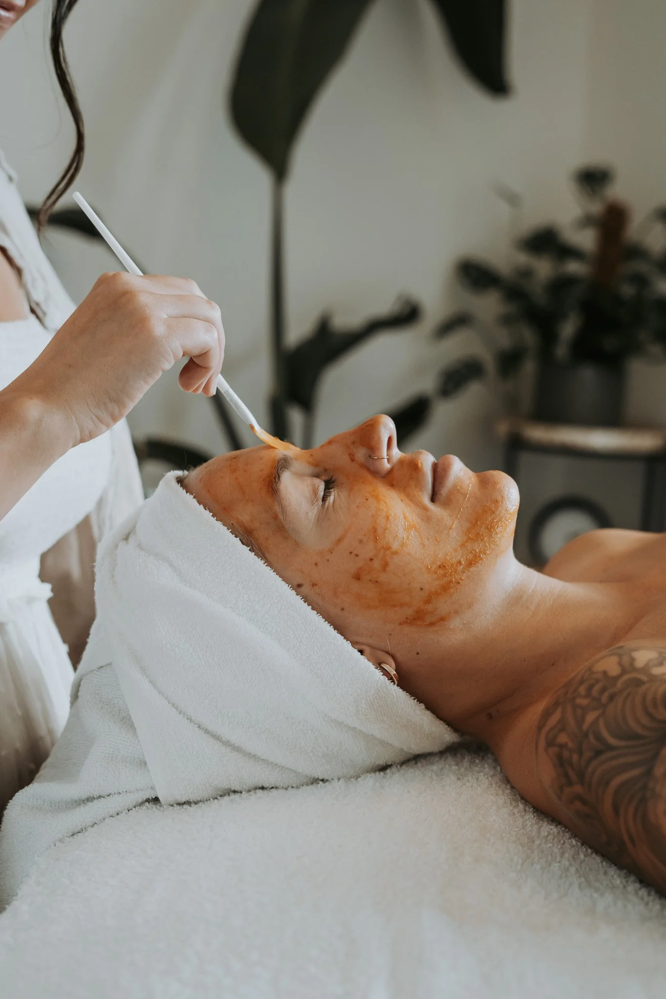 Person receiving a facial treatment with a facial mask, lying on a spa bed with a towel wrapped around their head.