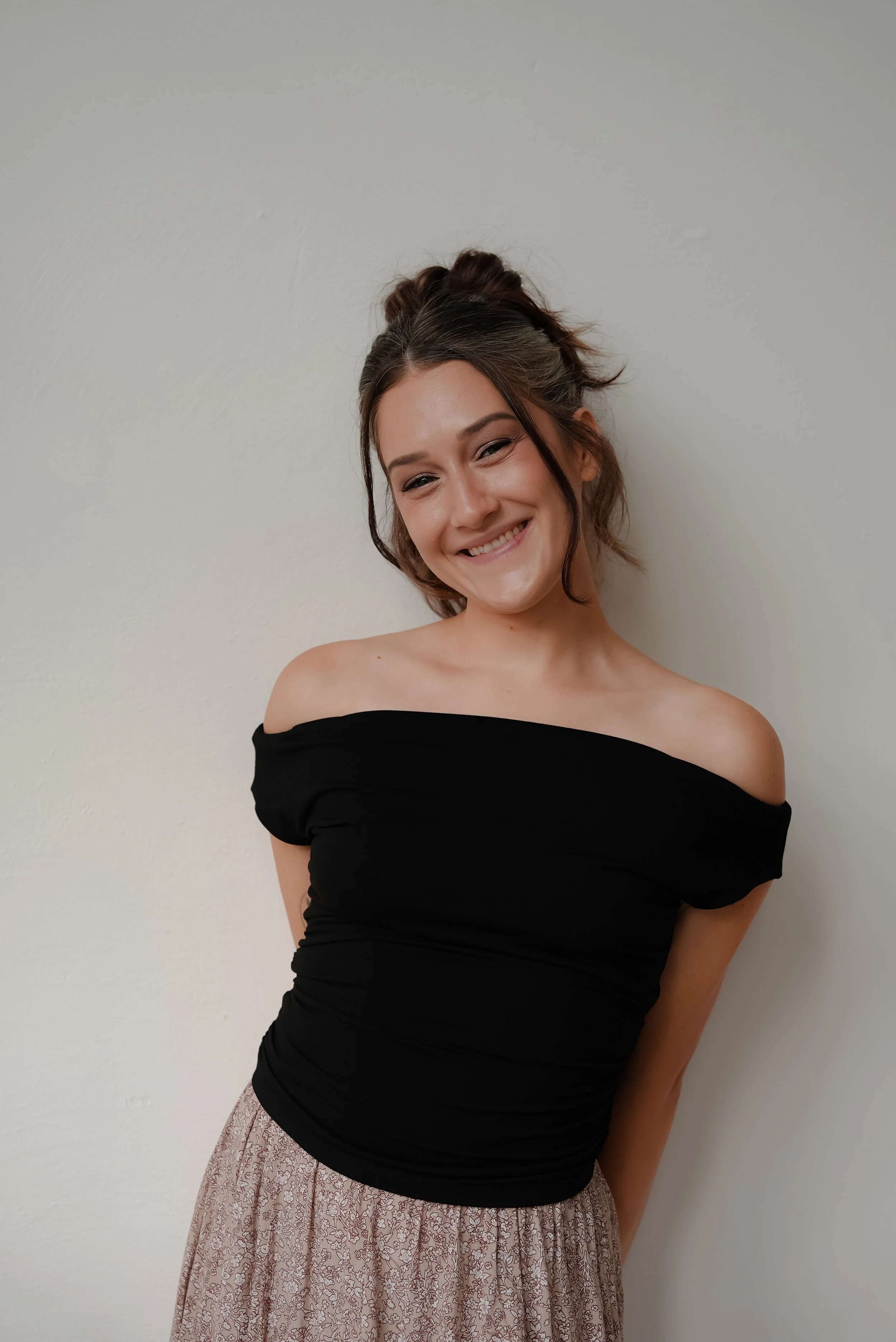 A young woman with dark hair in a bun, smiling, wearing an off-the-shoulder black top and a long patterned skirt, standing against a plain white wall.