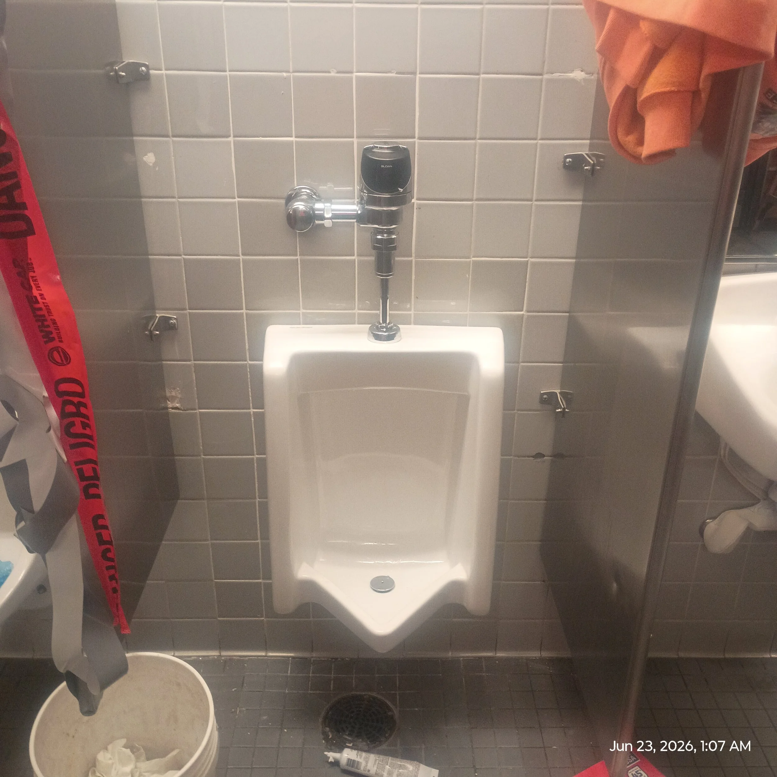 A bathroom stall with gray tiled walls and a small white sink. There is a black soap dispenser mounted on the wall above the sink, and a mirror partially visible on the right side. On the left, a red caution tape is hanging, and a white trash bin with crumpled tissue paper is in front of the stall. The timestamp indicates the photo was taken on June 23, 2026, at 1:07 AM.