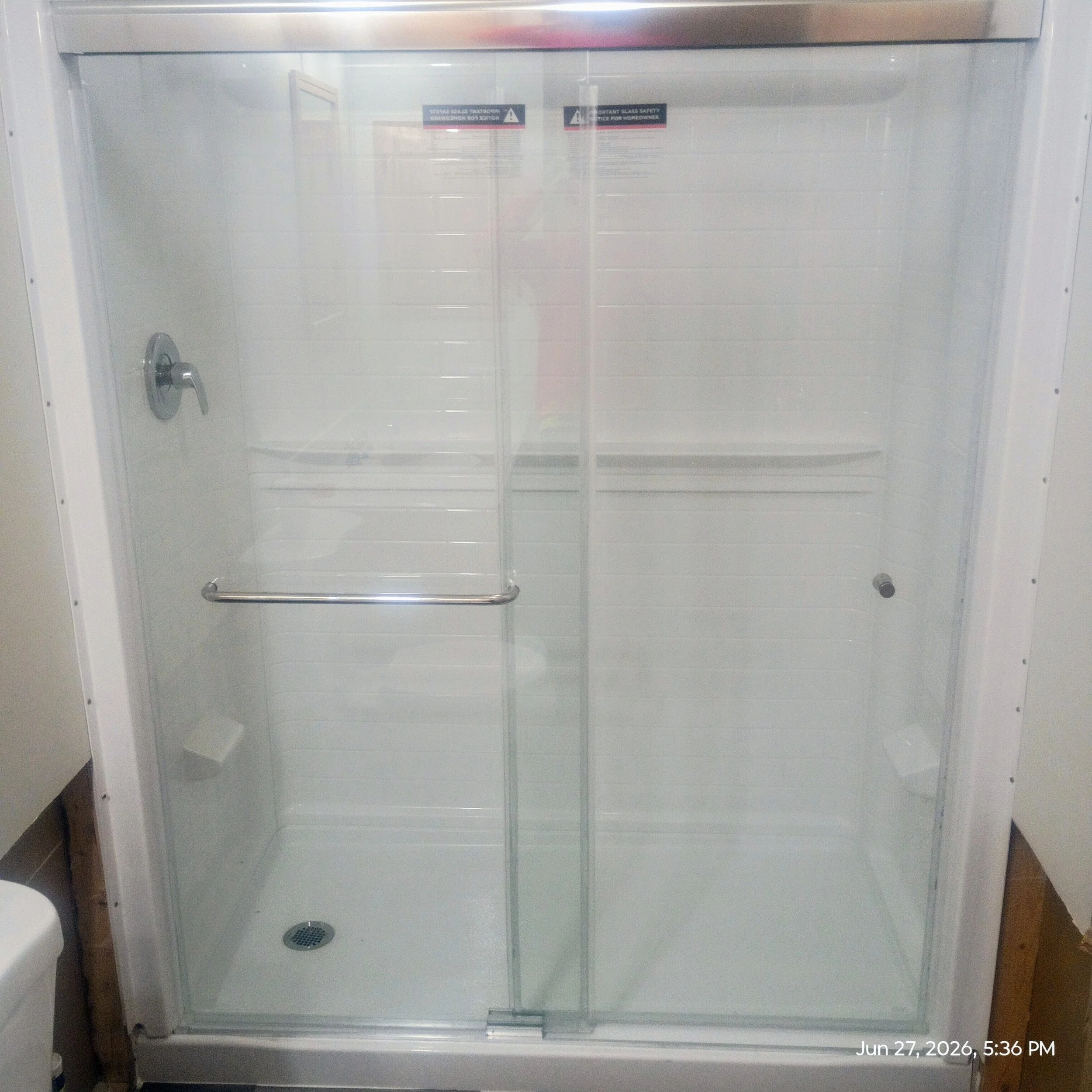Empty glass shower enclosure with a metal handle, drain at the bottom, and a timestamp in the lower right corner, June 27, 2026, 5:36 PM.