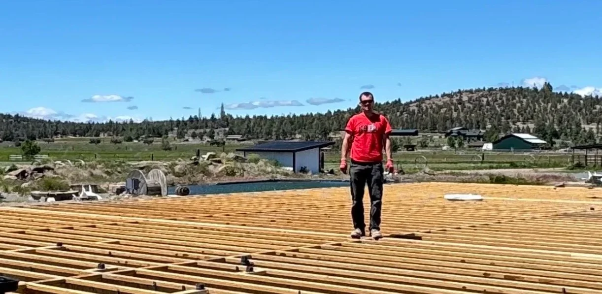 Person walking on a wooden construction site outdoors with a rural landscape and hills in the background.