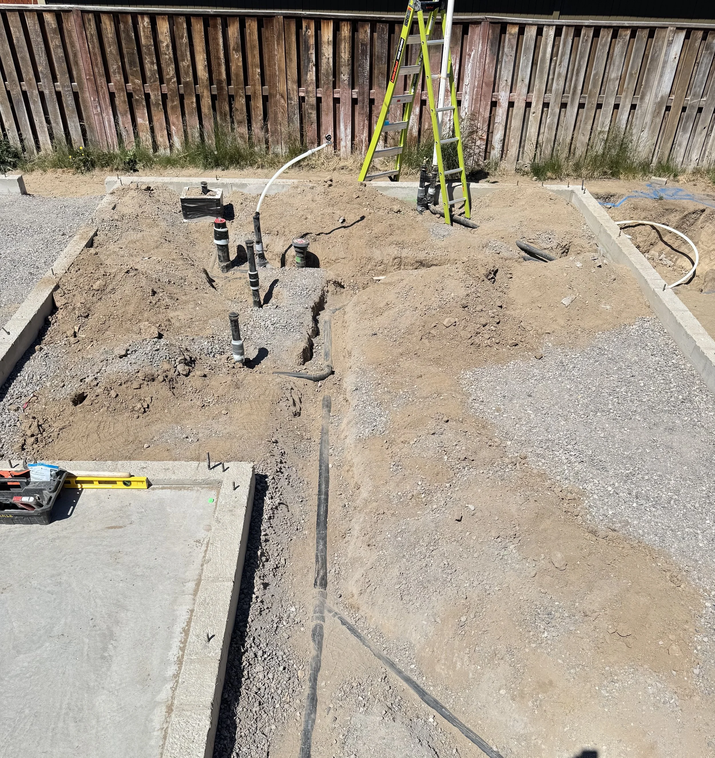 Construction site with dug trenches, piping, and a yellow ladder next to a wooden fence.