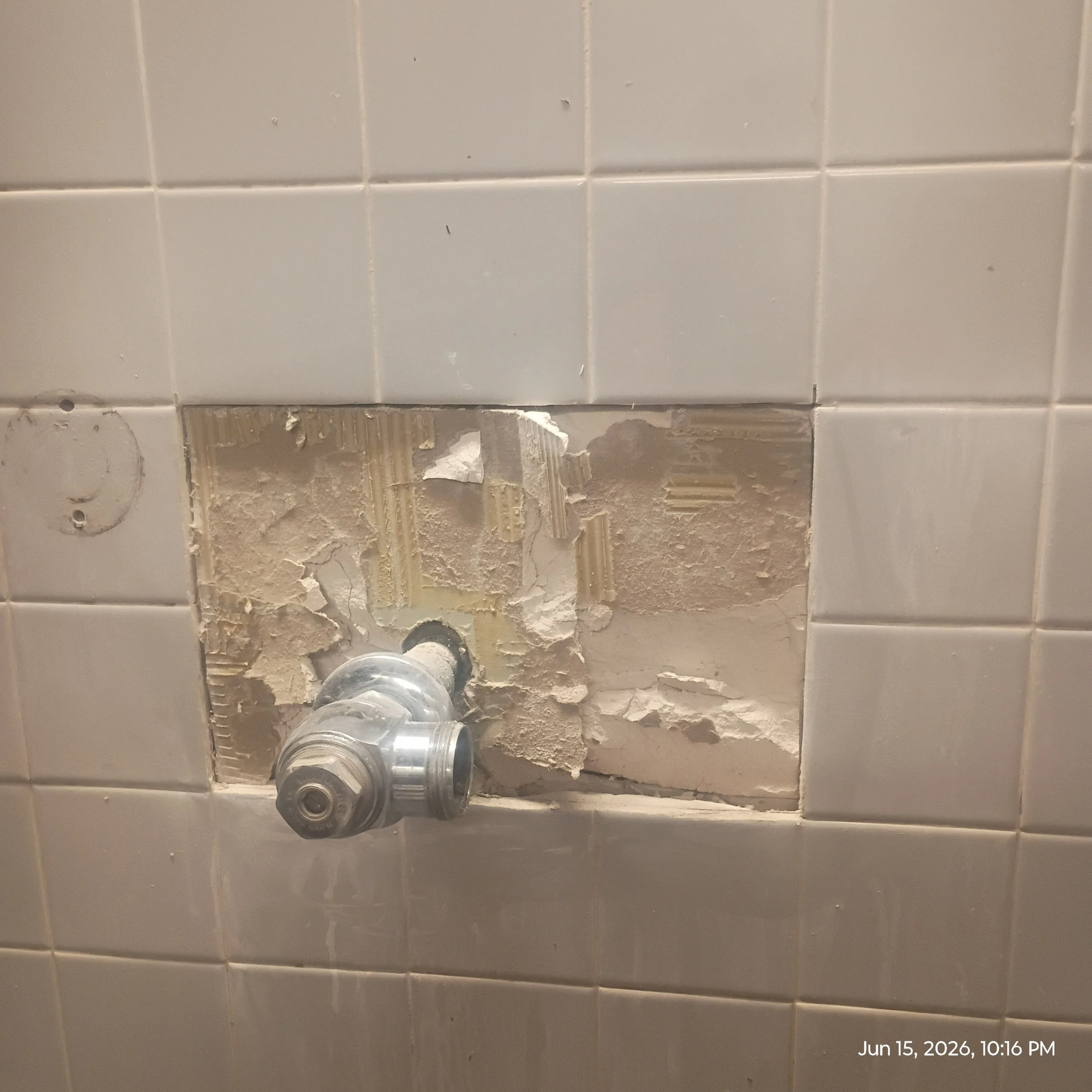 Broken and missing bathroom tiles with exposed wall underneath and a partially visible silver pipe.