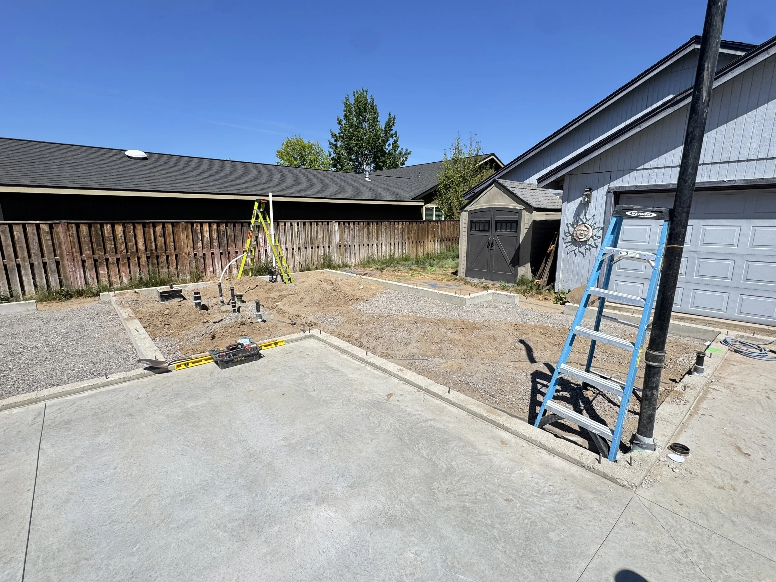 A backyard under construction with exposed soil and several pipes sticking out, a wooden fence, a gray shed, a ladder, and part of a house with a garage door, all under a clear blue sky.