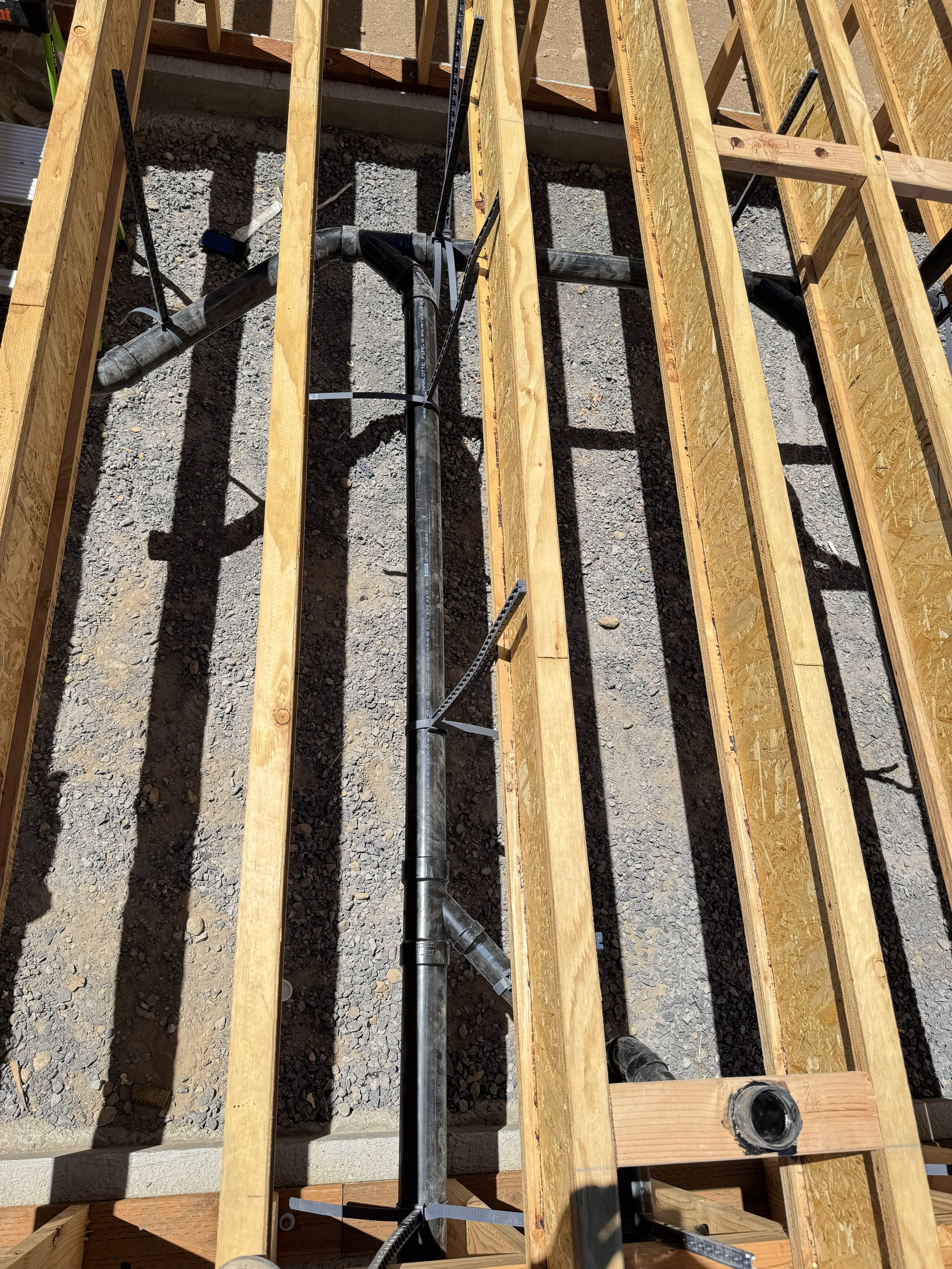 View of a construction site showing wooden framing for a floor, with black plumbing pipes installed underneath, sunlight casting shadows, and gravel ground visible.