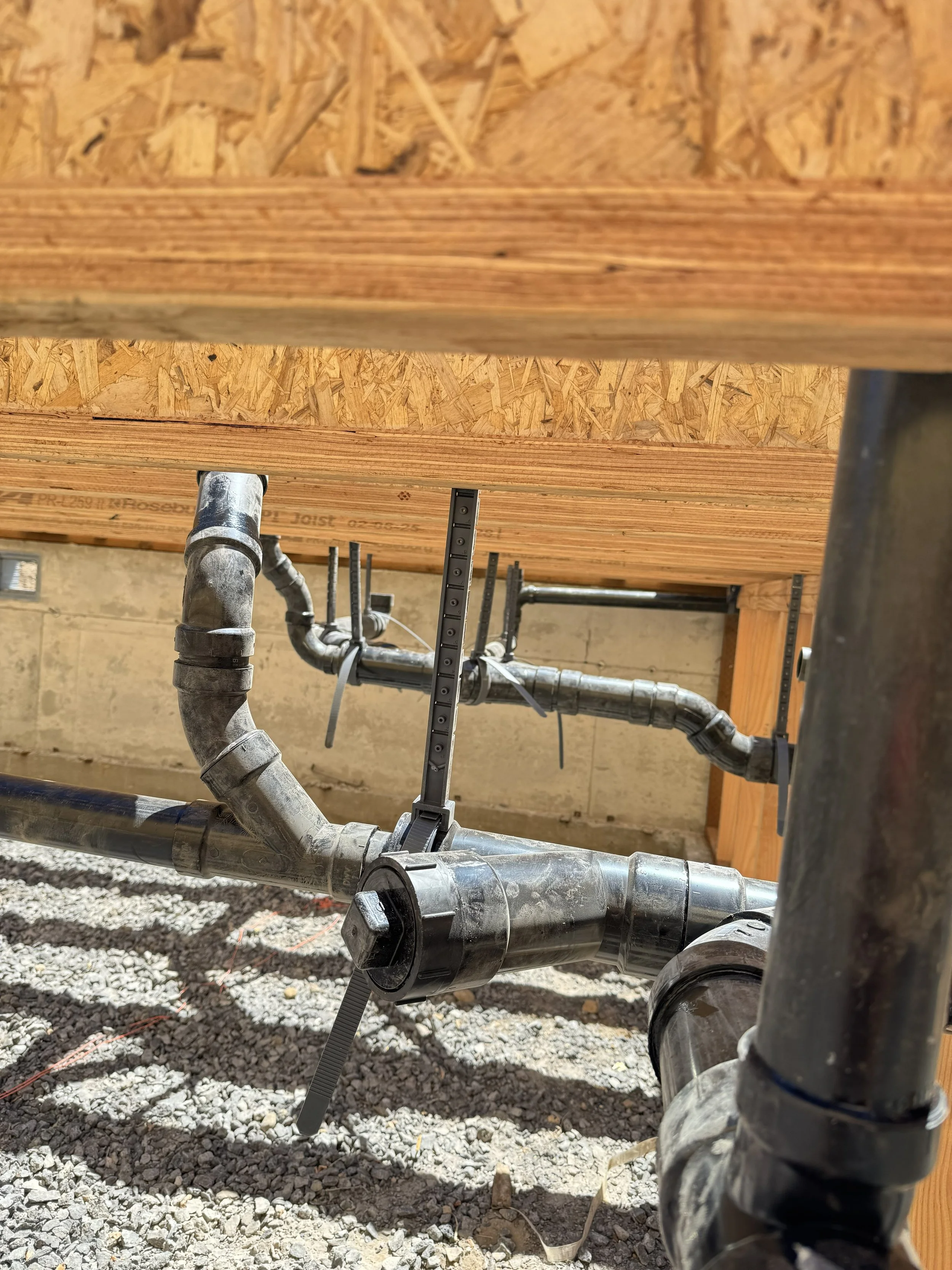 The underside of a building’s wooden floor structure with visible plumbing pipes and gravel ground.