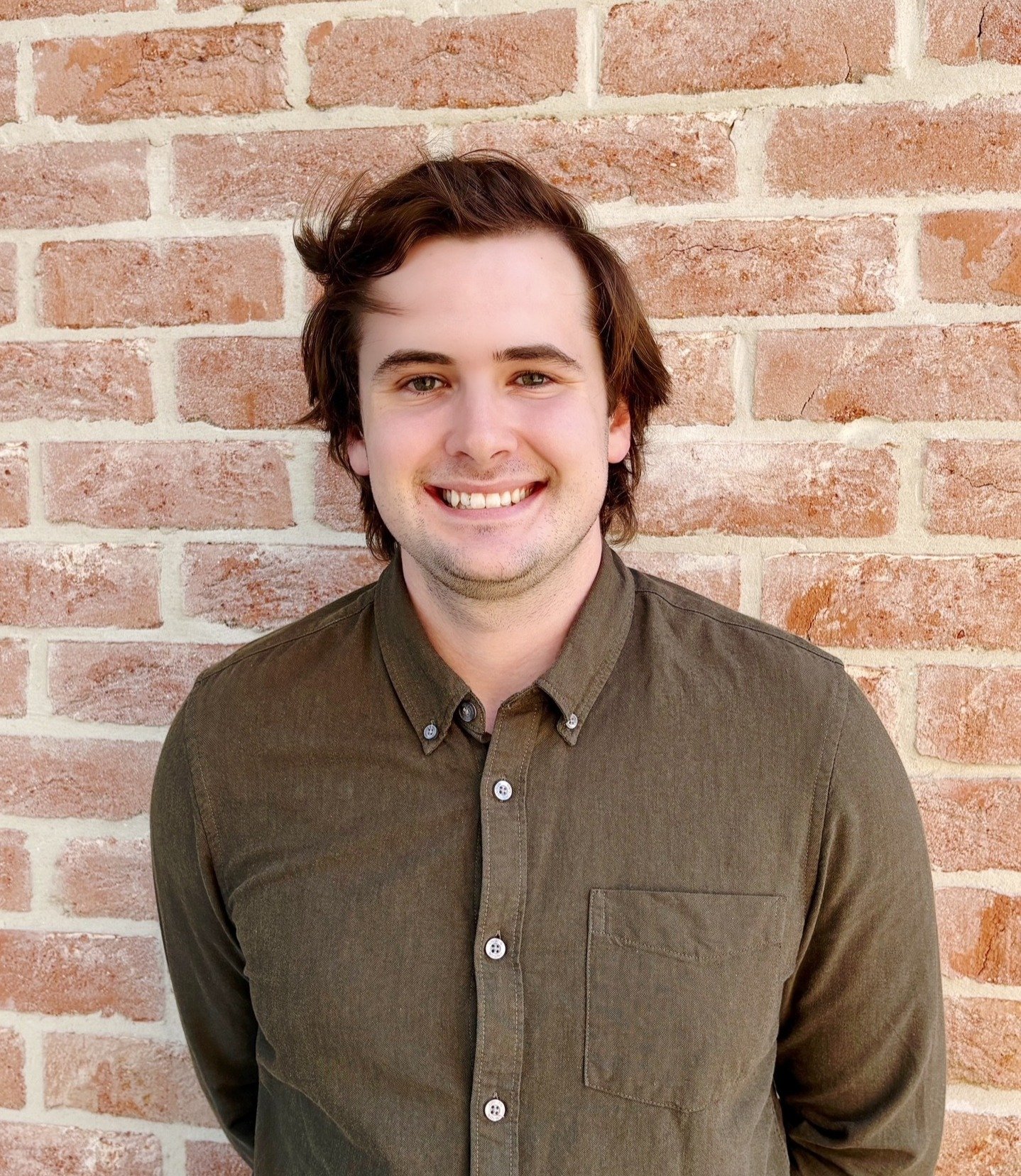 A young man with brown hair and green eyes smiling, wearing a brown button-up shirt, standing in front of a red brick wall.