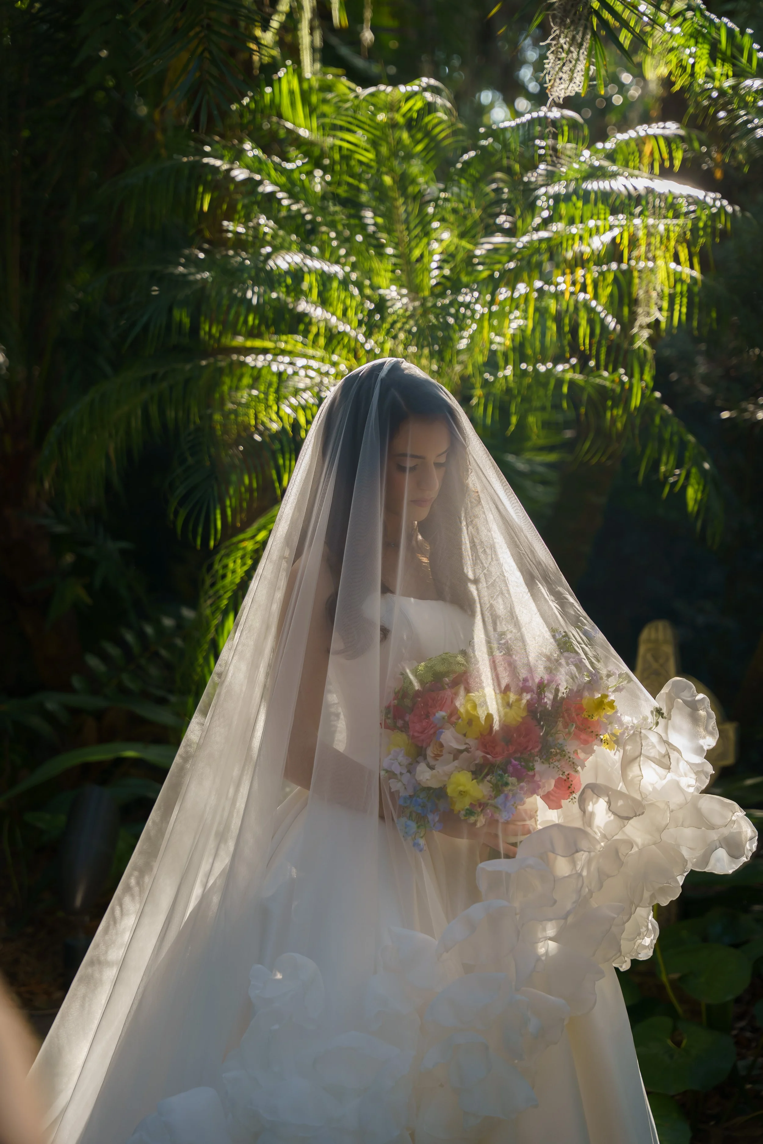 A woman in a wedding dress with a veil holding a bouquet of colorful flowers, standing outdoors with lush green foliage in the background.