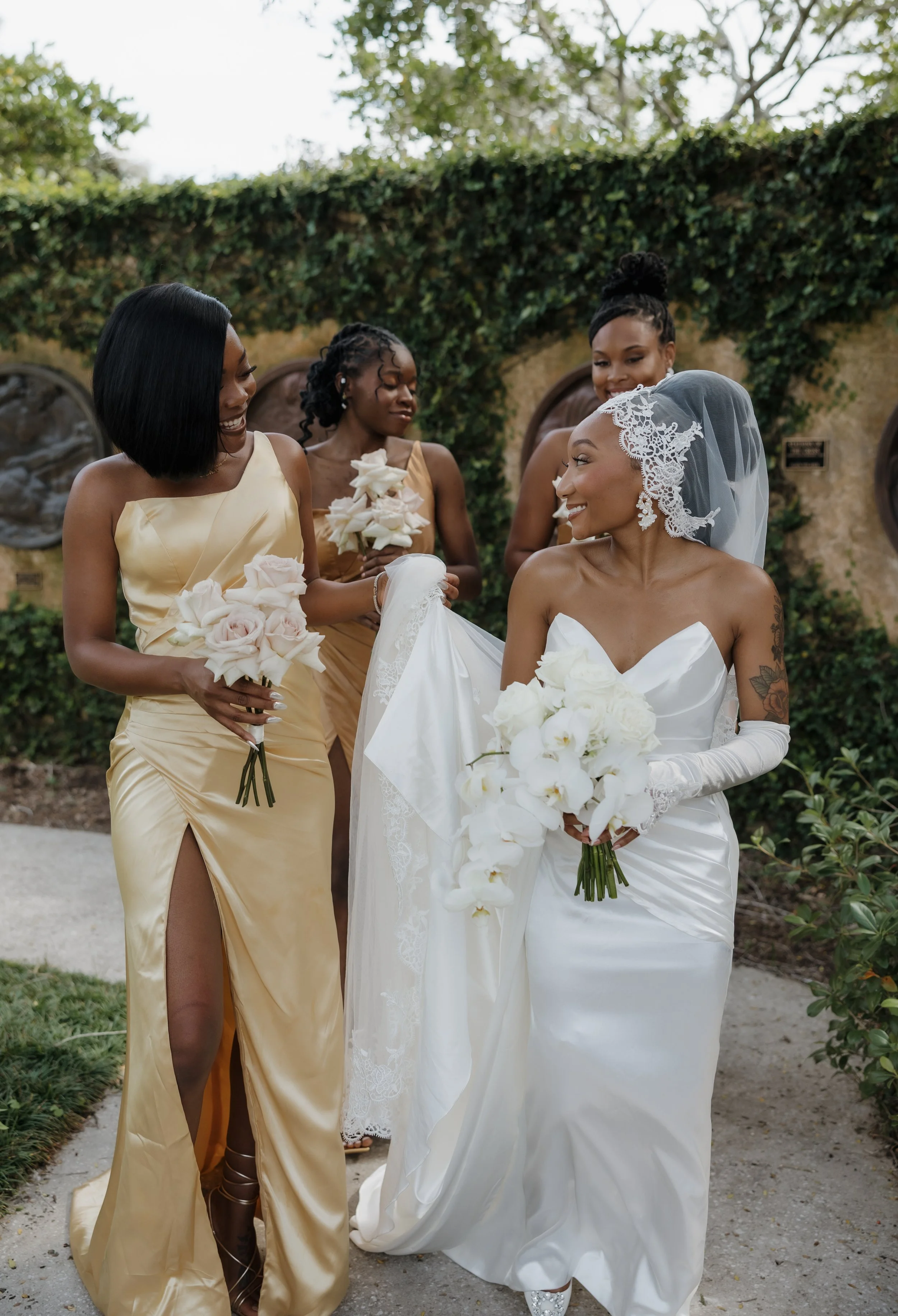 A bride in a white wedding dress and lace veil holding white orchids, smiling at a woman in a gold gown, surrounded by three women in elegant dresses, outdoors with green foliage in the background.