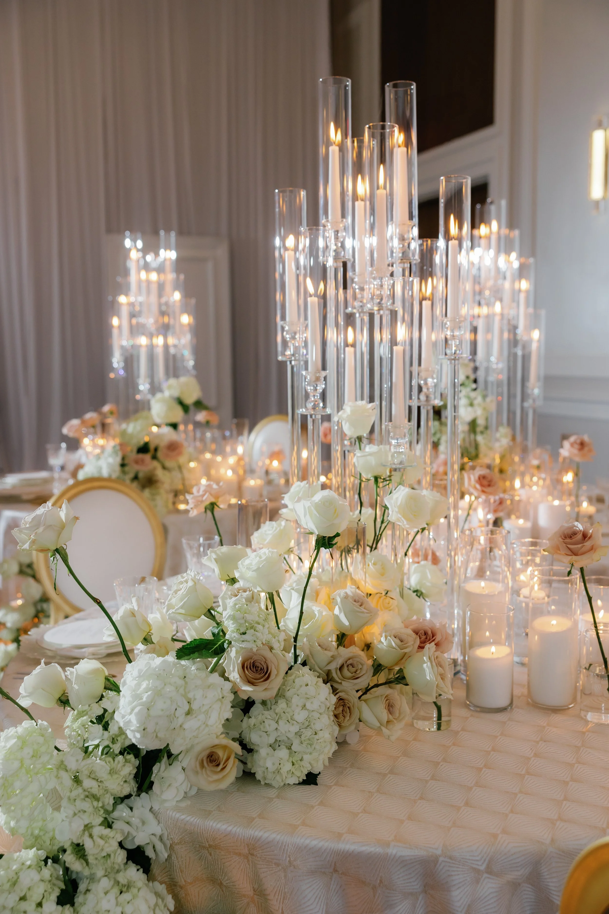 Elegant wedding reception table decorated with white and blush roses, hydrangeas, tall glass candle holders with lit candles, and white pillar candles in glass holders. The background features cream-colored drapes and a wall-mounted light.
