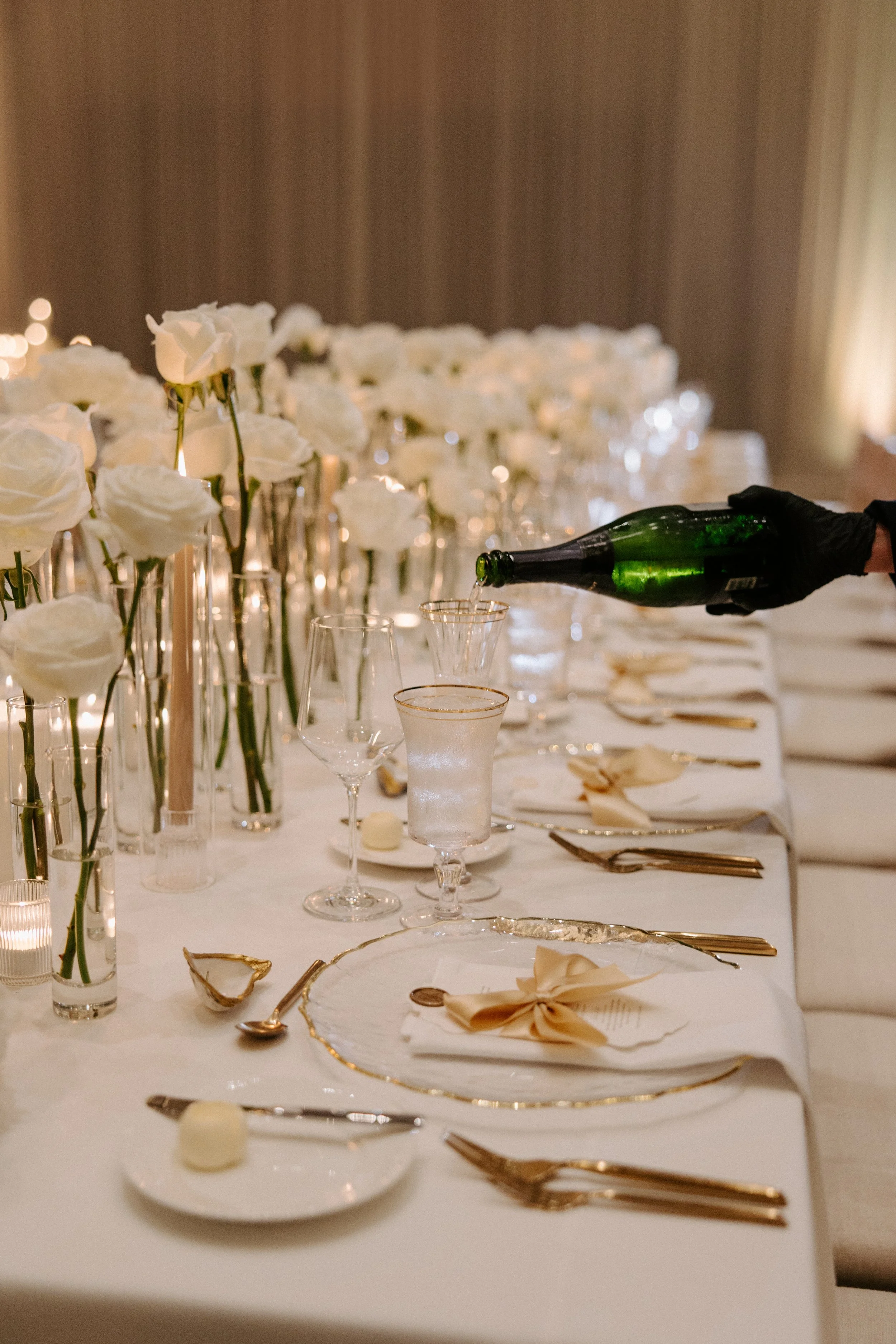 A formal dining table decorated with white flowers, gold-rimmed plates, gold cutlery, and glasses. Someone is pouring champagne into a glass. The table is set with folded napkins and small decorative items.