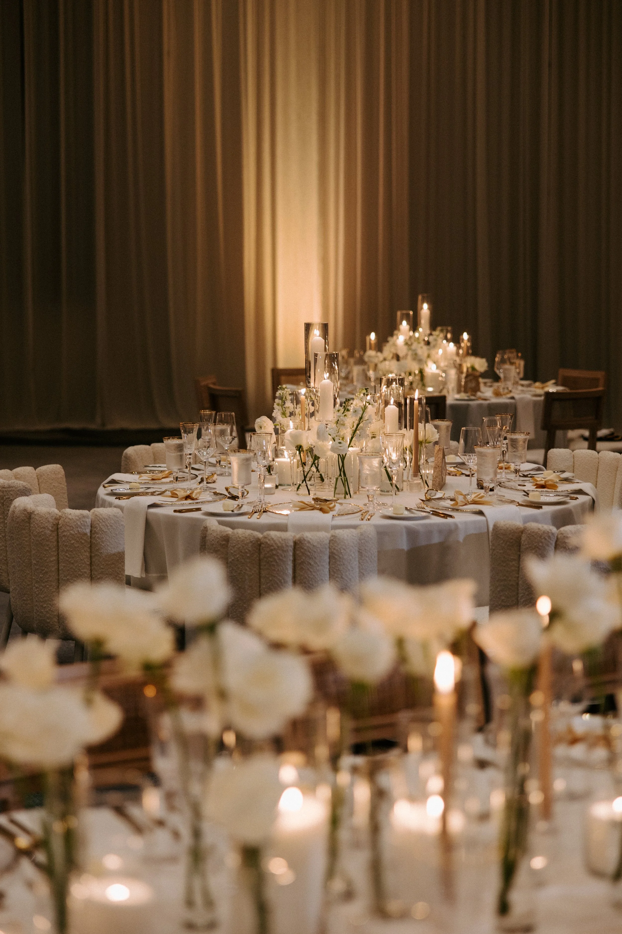 Elegant banquet table decorated with white flowers, candles, and glassware in a softly lit reception hall with dark curtains.