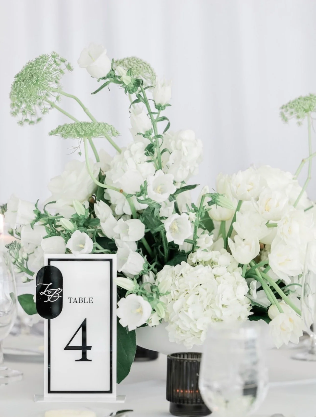 White wedding reception flower arrangement on a table with a black table number sign showing 'Table 4'. The flowers are predominantly white with some greenery, possibly at a formal event or wedding.