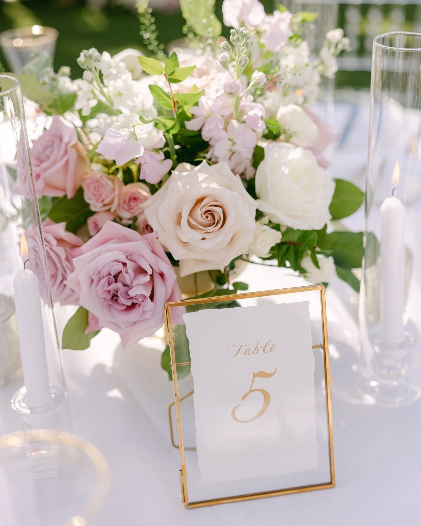 A floral centerpiece with white and pink roses, and light pink and white flowers, surrounded by glass candle holders on a white table with a table number sign reading 'Table 5.'