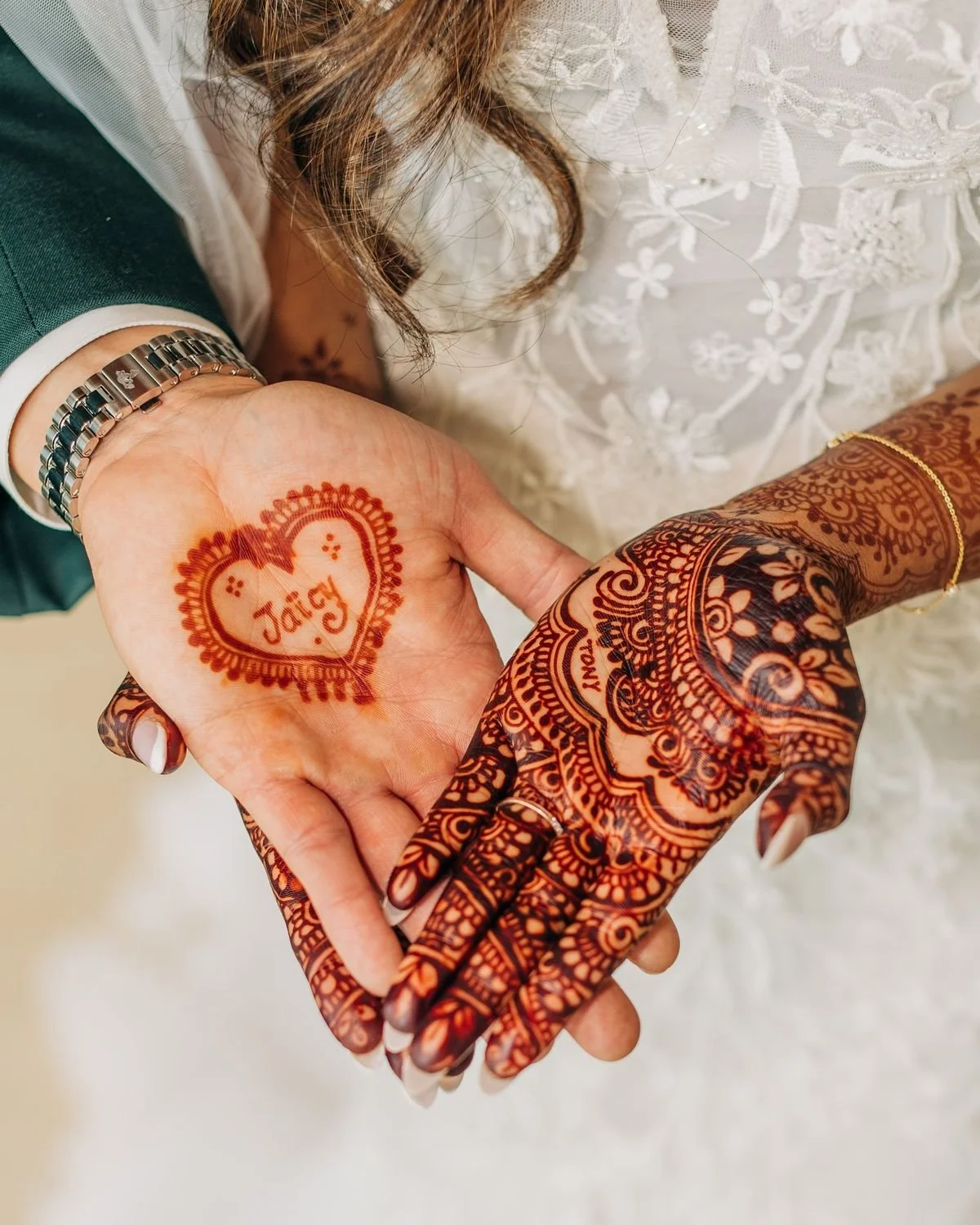 Close-up of a newlywed couple holding hands, displaying henna designs on their hands. The bride's hand has a heart-shaped mehndi pattern with the name 'Jay' written inside. The groom's hand features intricate henna art, with visible rings and jewelry, in front of a white lace wedding dress.