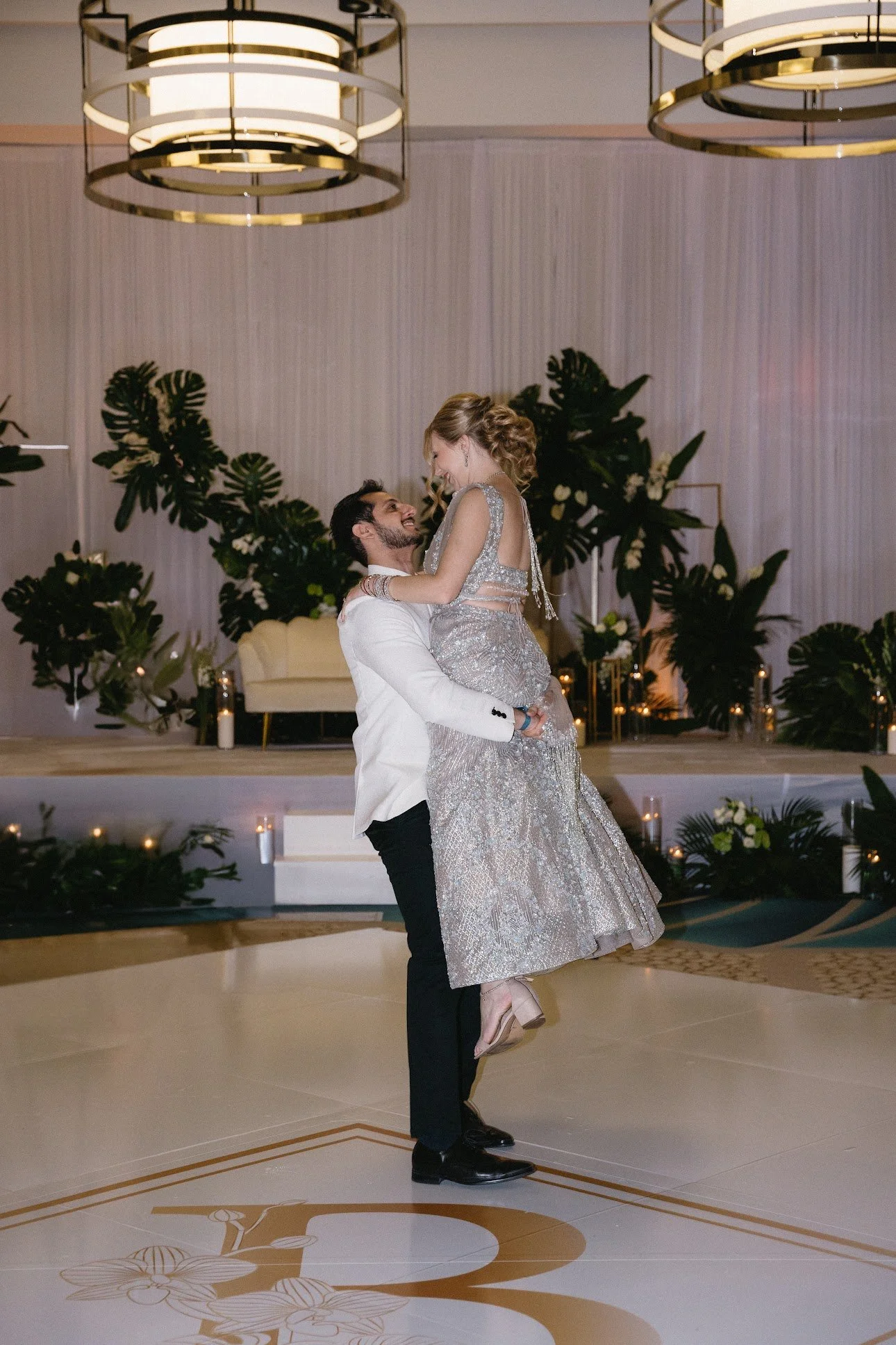 A man in a white tuxedo lifts a woman in a silver gown during a dance at a formal event with elegant lighting and lush green plants in the background.