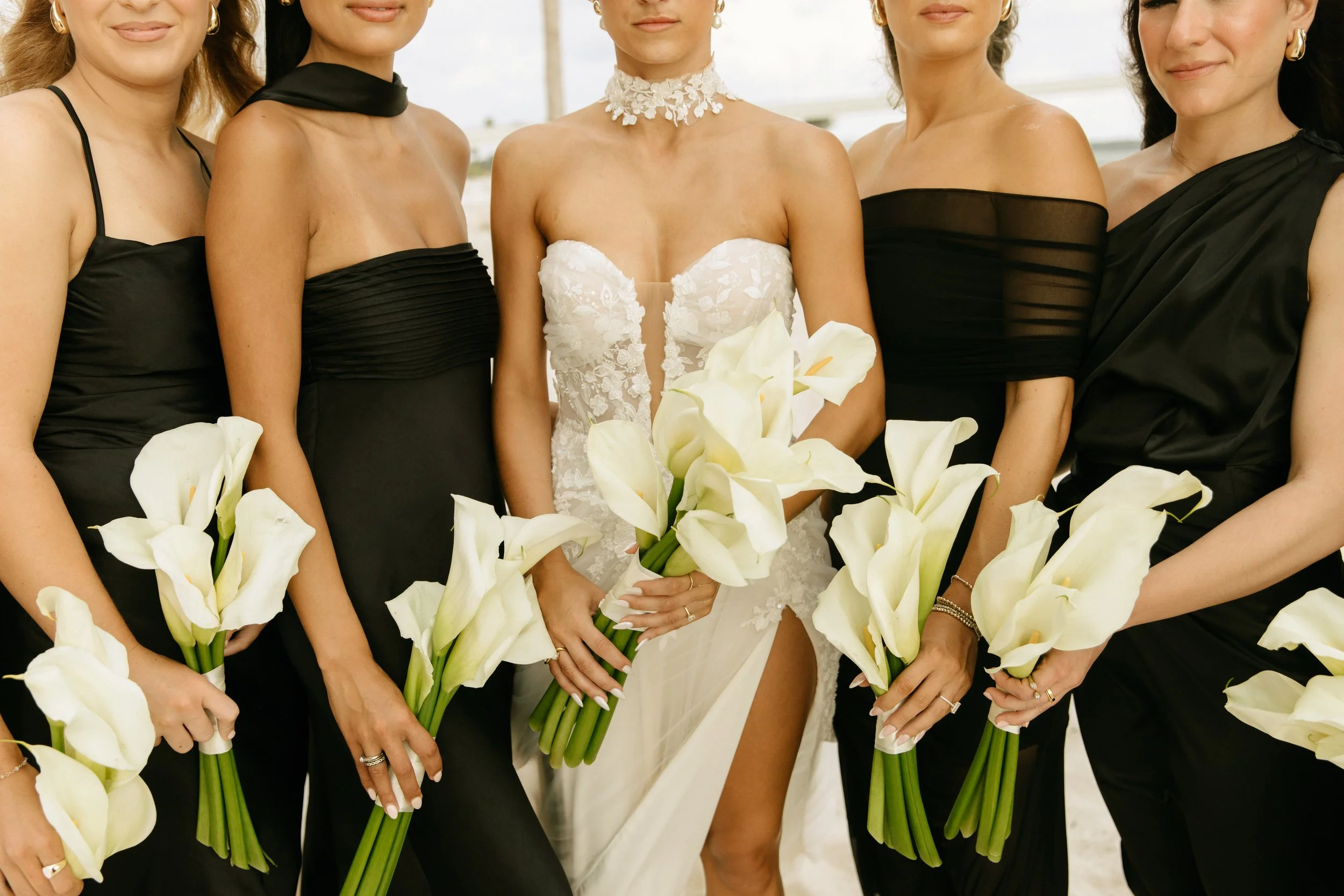 A group of women in black dresses standing together holding white calla lilies, with one woman in a white bridal gown in the center.