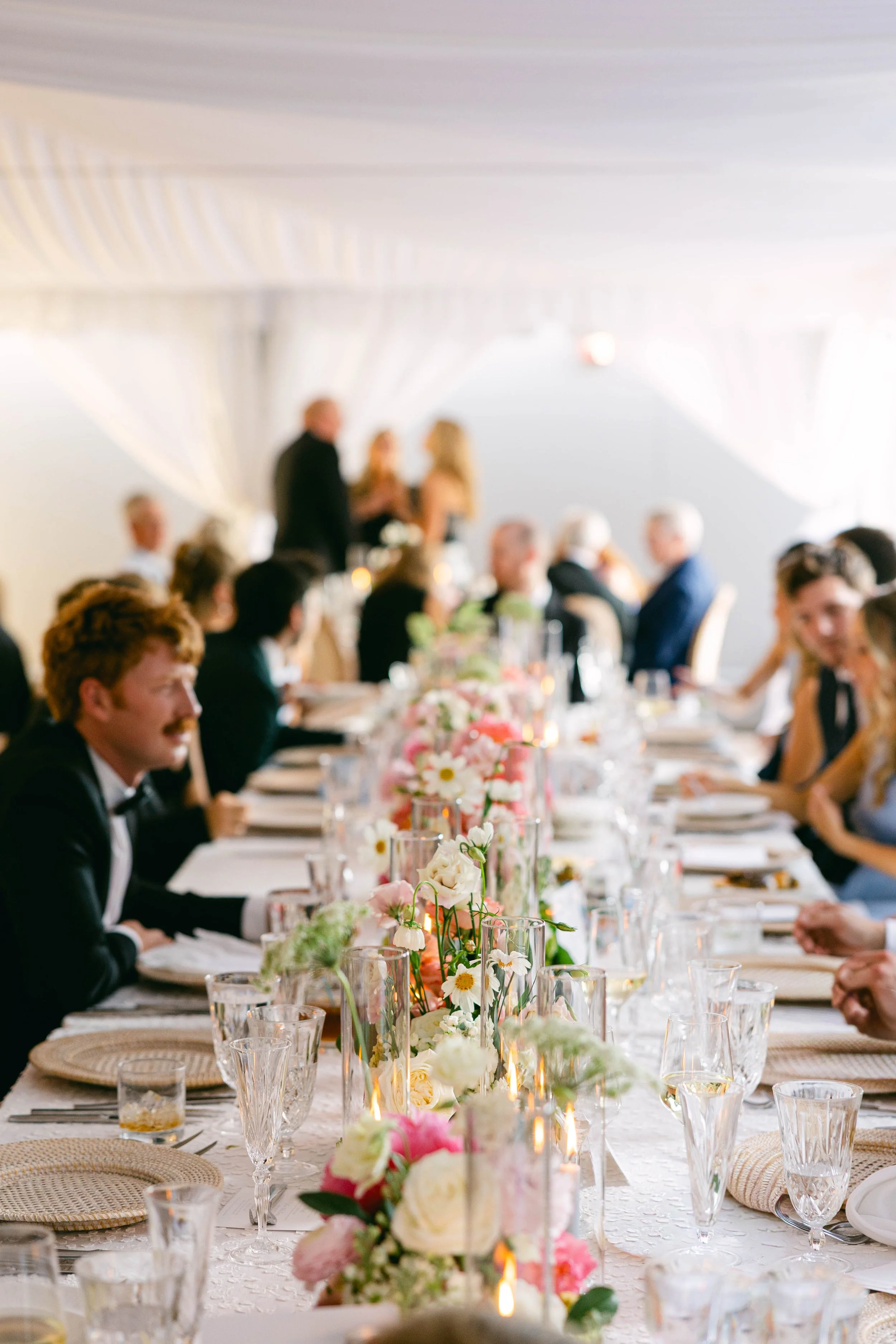 A wedding reception with a long decorated table featuring floral centerpieces, candles, glassware, and plates, with guests seated and a couple standing and talking in the background.