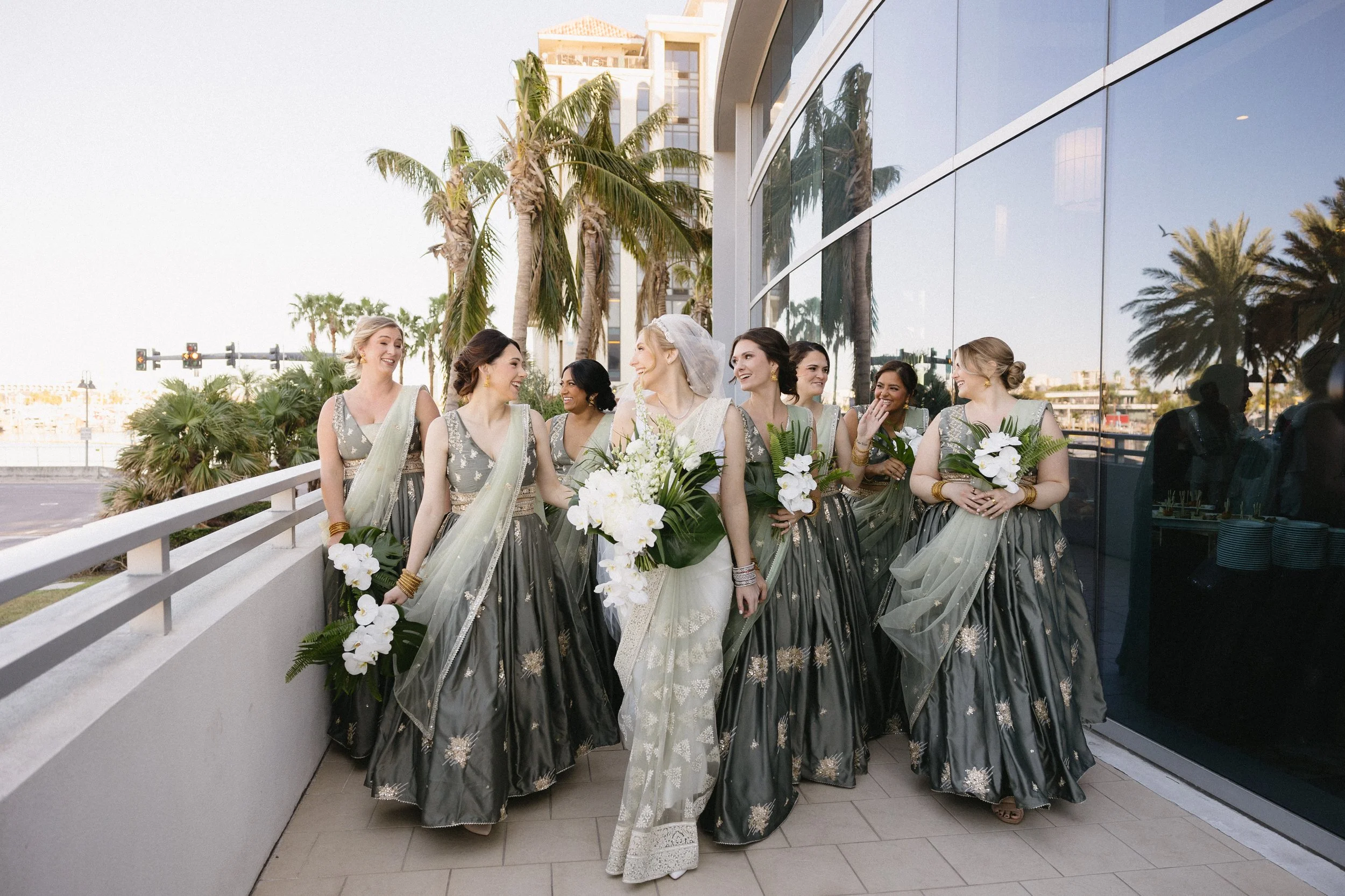 Group of women in traditional Indian sarees and jewelry, holding flowers, standing outdoors near tall palm trees and reflecting glass building, during a wedding or festive event.